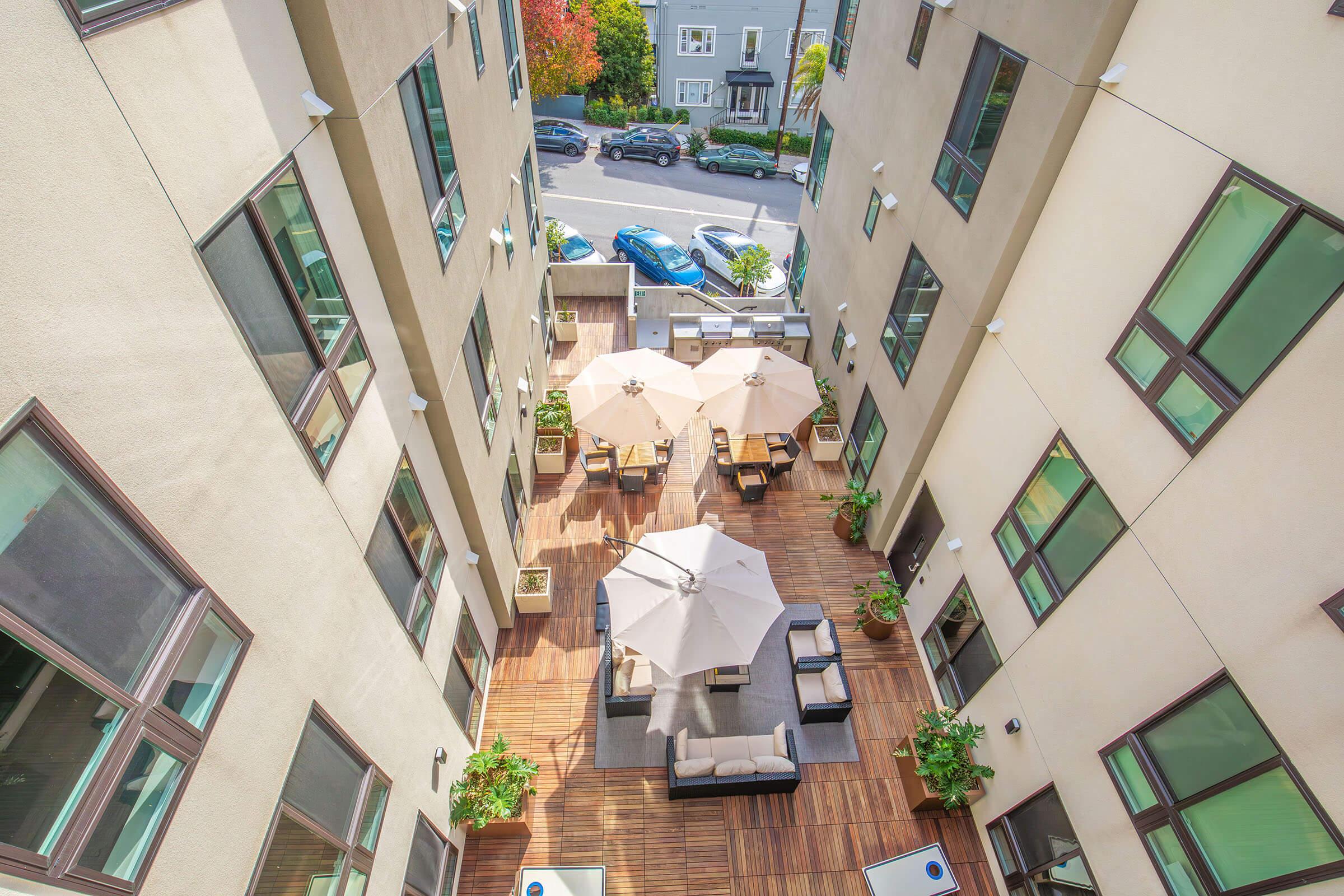 A view of a modern outdoor courtyard surrounded by tall buildings, featuring multiple patio tables under large umbrellas. The area has wooden decking, comfortable seating, and potted plants, with cars parked on the street in the background. Sunlight enhances the inviting atmosphere.