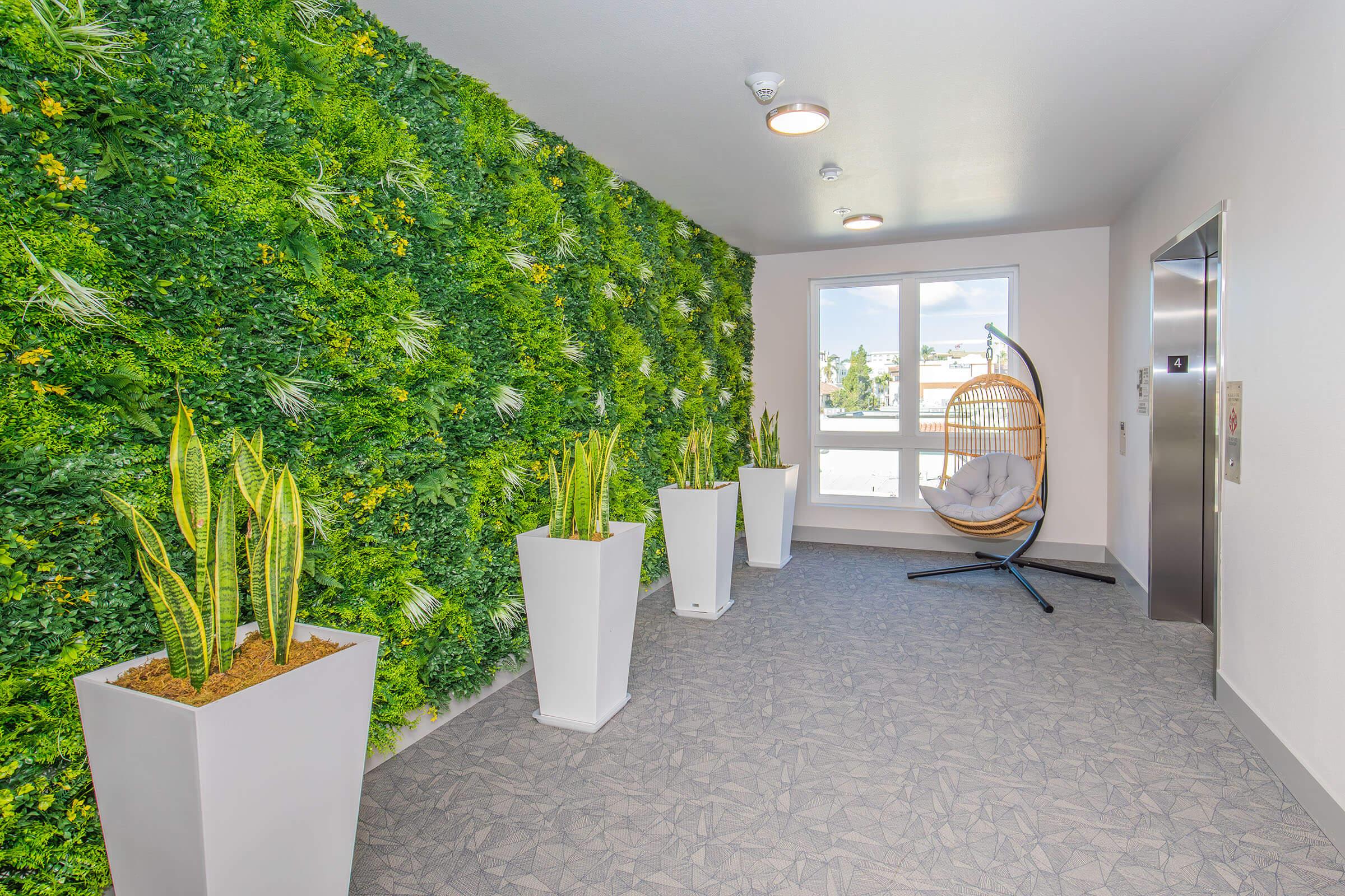 A modern hallway featuring a vibrant green living wall with various plants, flanked by tall, white planters containing snake plants. A cozy hanging chair is positioned near a large window, and an elevator is visible on the right side. The overall design is bright and inviting.