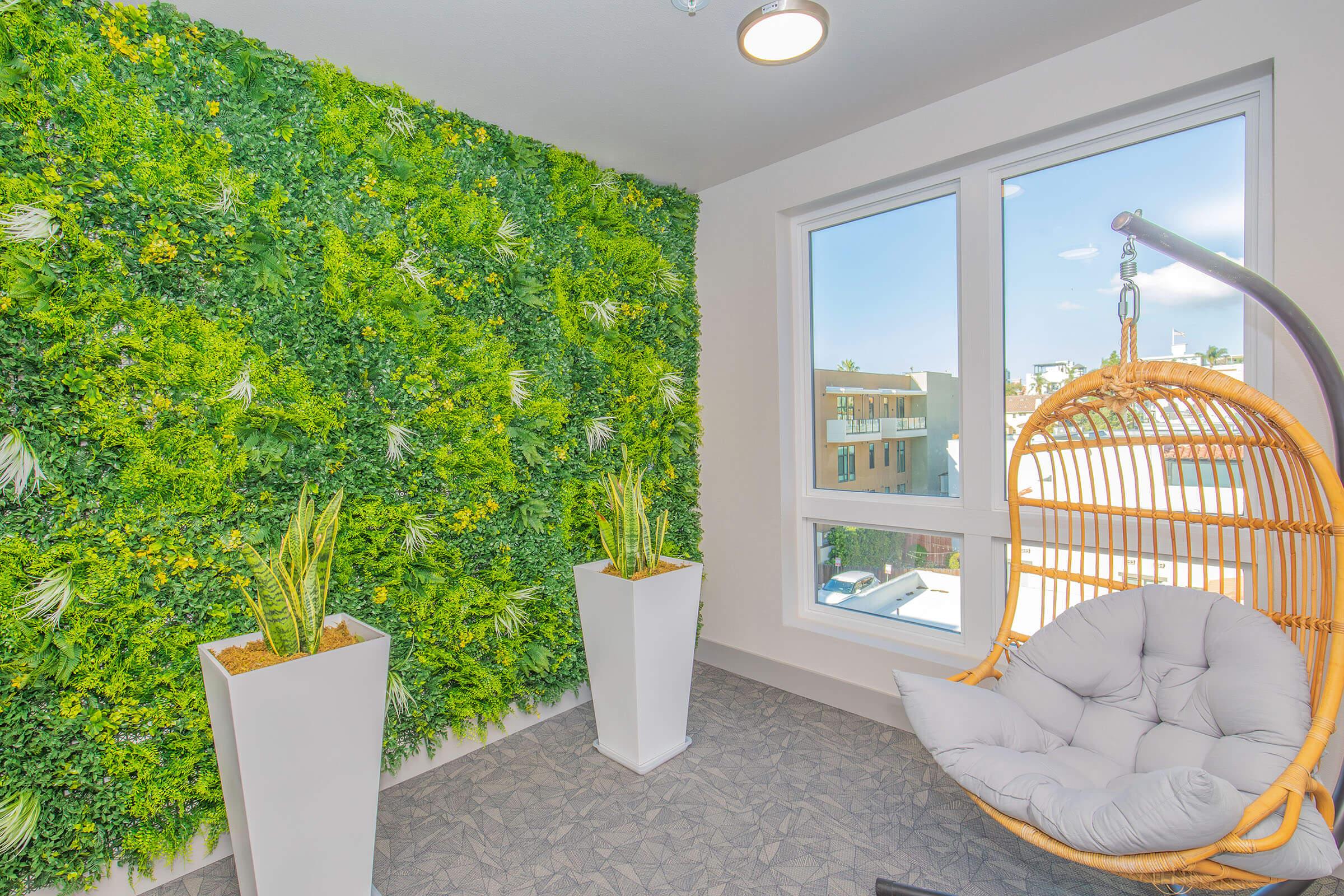A cozy indoor space featuring a vibrant green artificial plant wall, two potted plants, and a hanging rattan chair with a plush cushion. Large windows allow natural light to fill the room, providing a view of the outdoors. The flooring is a textured gray.