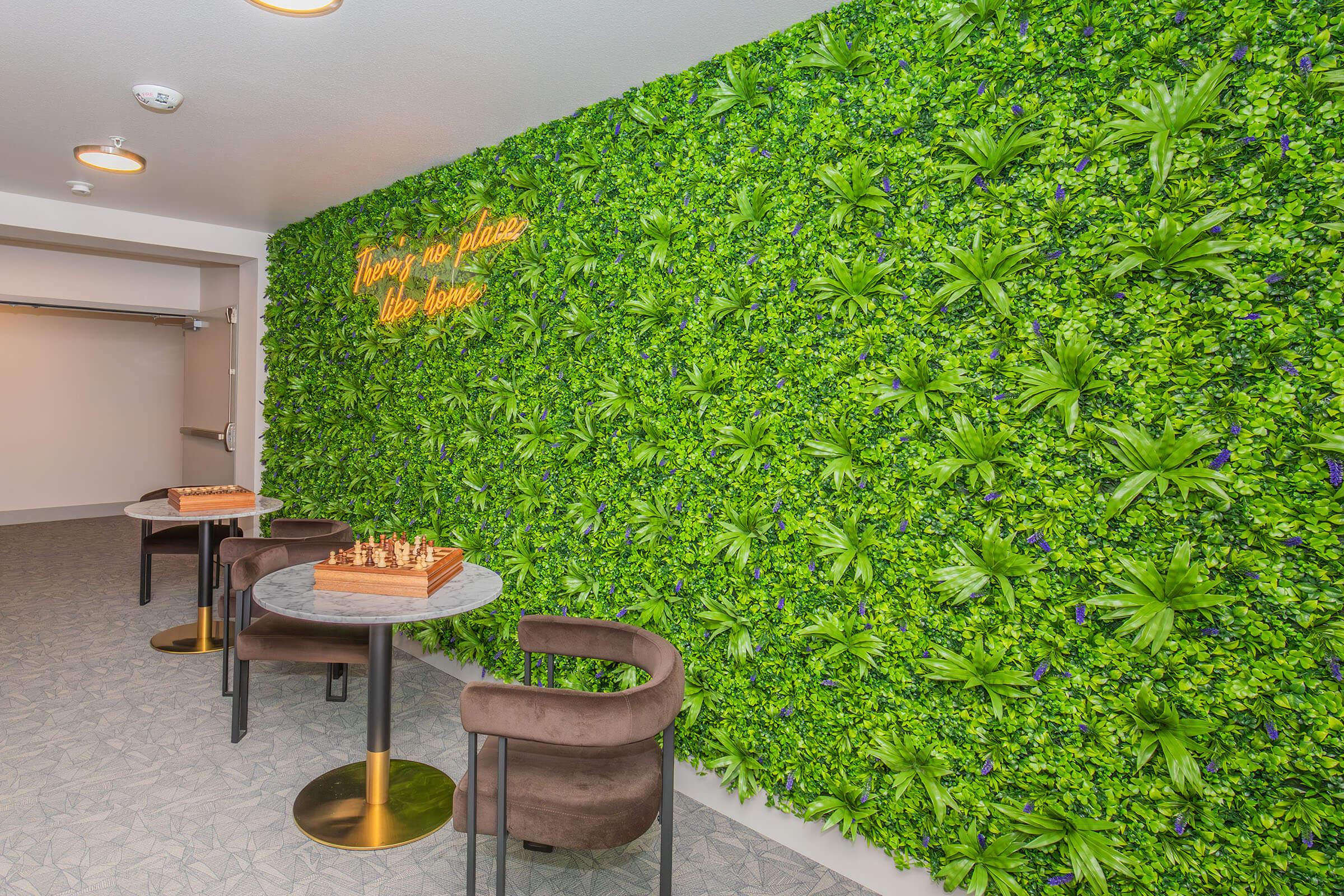 A vibrant green wall covered in artificial foliage, featuring small purple flowers. In front, there is a round marble table with a wooden chess set. Two modern, dark upholstered chairs are positioned nearby, creating a cozy and inviting atmosphere in a lobby or lounge area. Decorative lighting adds warmth to the space.