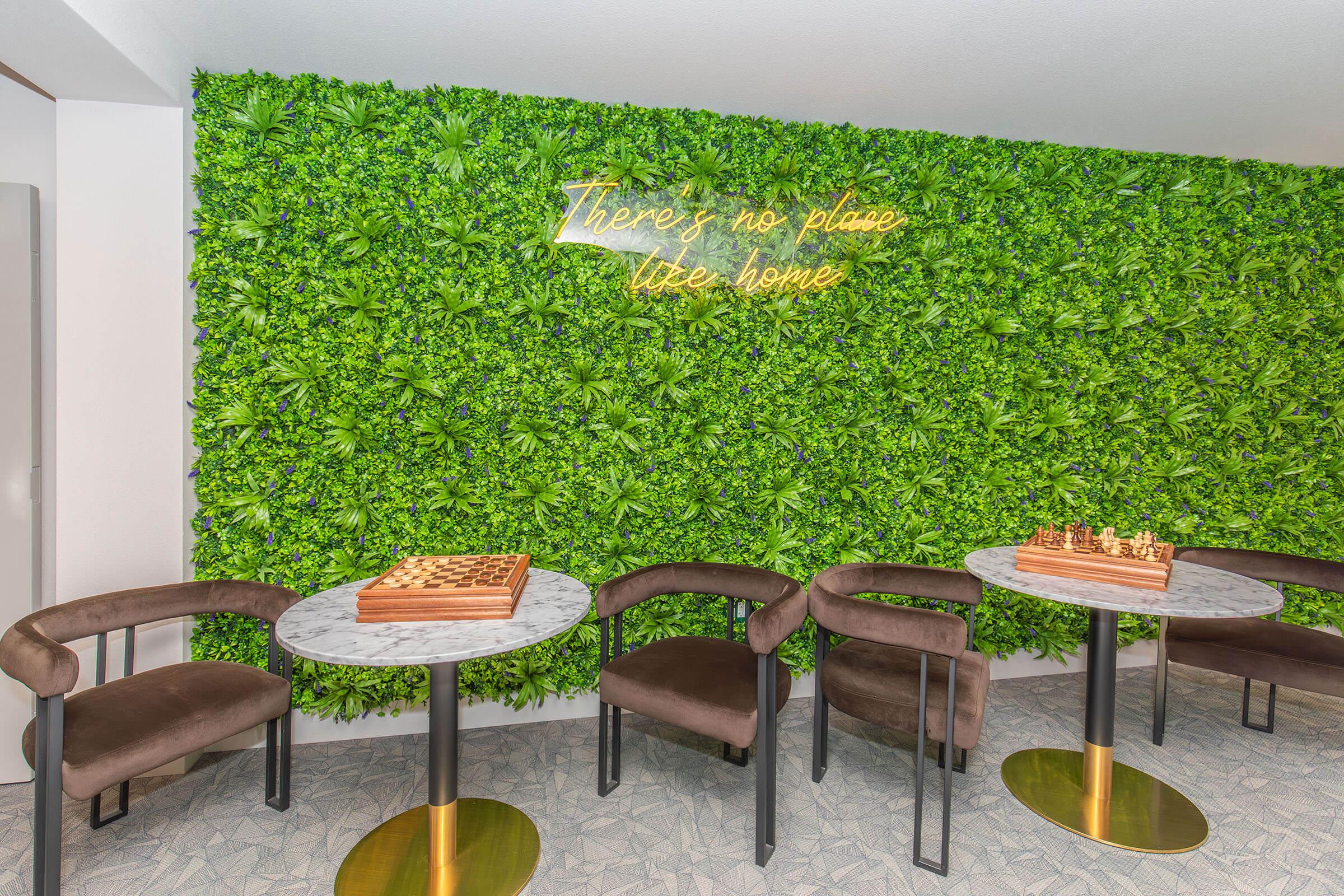 A cozy indoor area featuring a green wall of artificial plants, with a neon sign that reads "There's no place like home." There are two round marble-top tables with stylish black chairs, and a wooden chess set on one table, creating a welcoming and relaxed atmosphere.