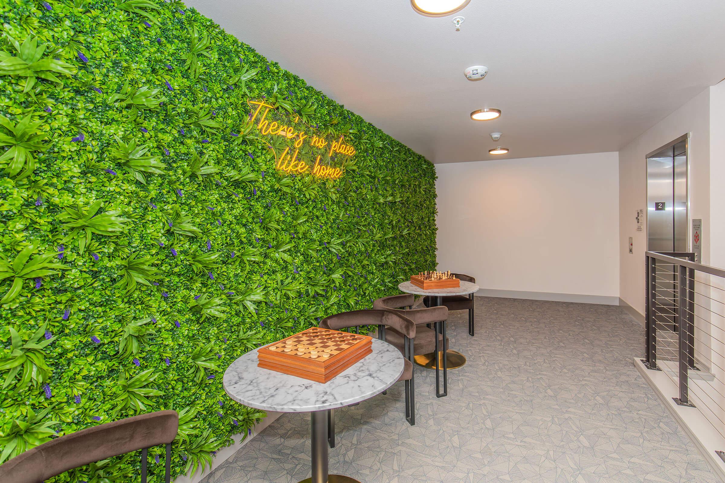 A cozy interior space featuring a green living wall with the quote "There's no place like home" illuminated in neon. There are two small marble-topped tables with cushioned chairs, and a chess set on one table, creating an inviting atmosphere for relaxation or socializing.