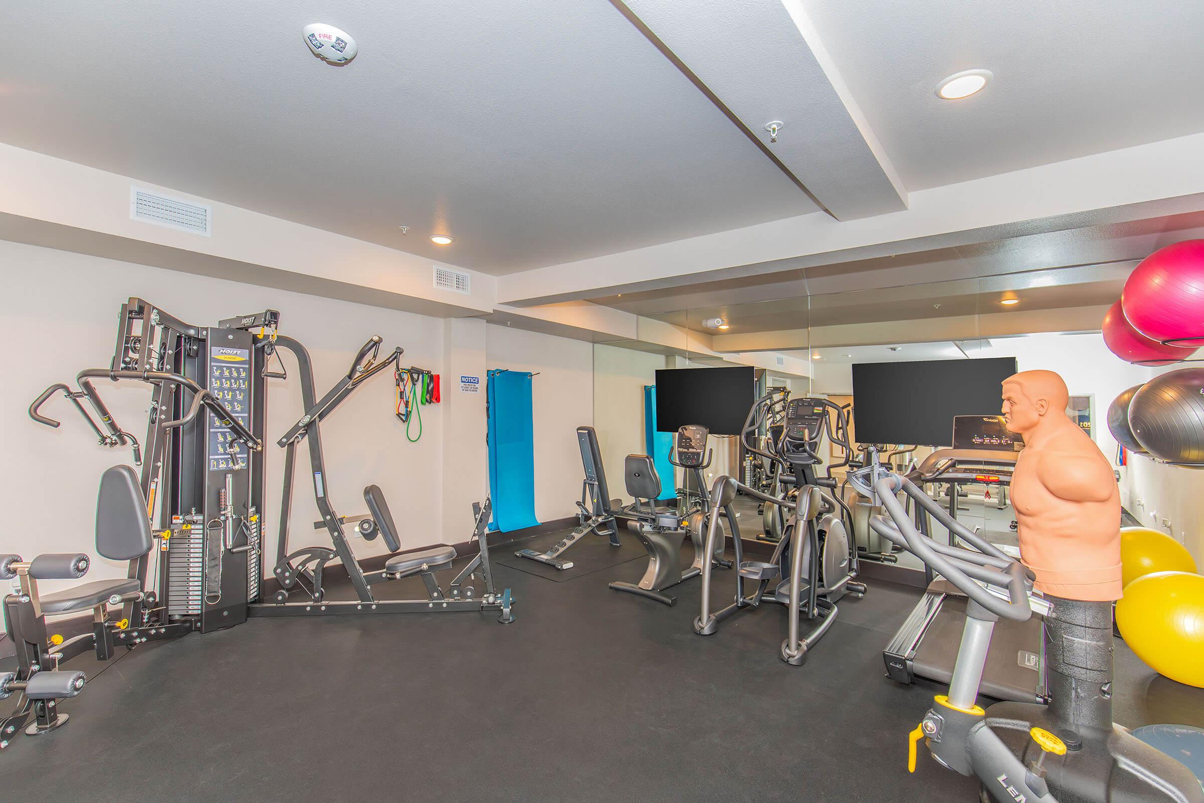 A modern gym space featuring various exercise equipment, including weight machines, treadmills, and stationary bicycles. There is a training dummy, fitness balls in different colors, and large mirrors reflecting the room. Bright, clean environment with a neutral color palette and good lighting.