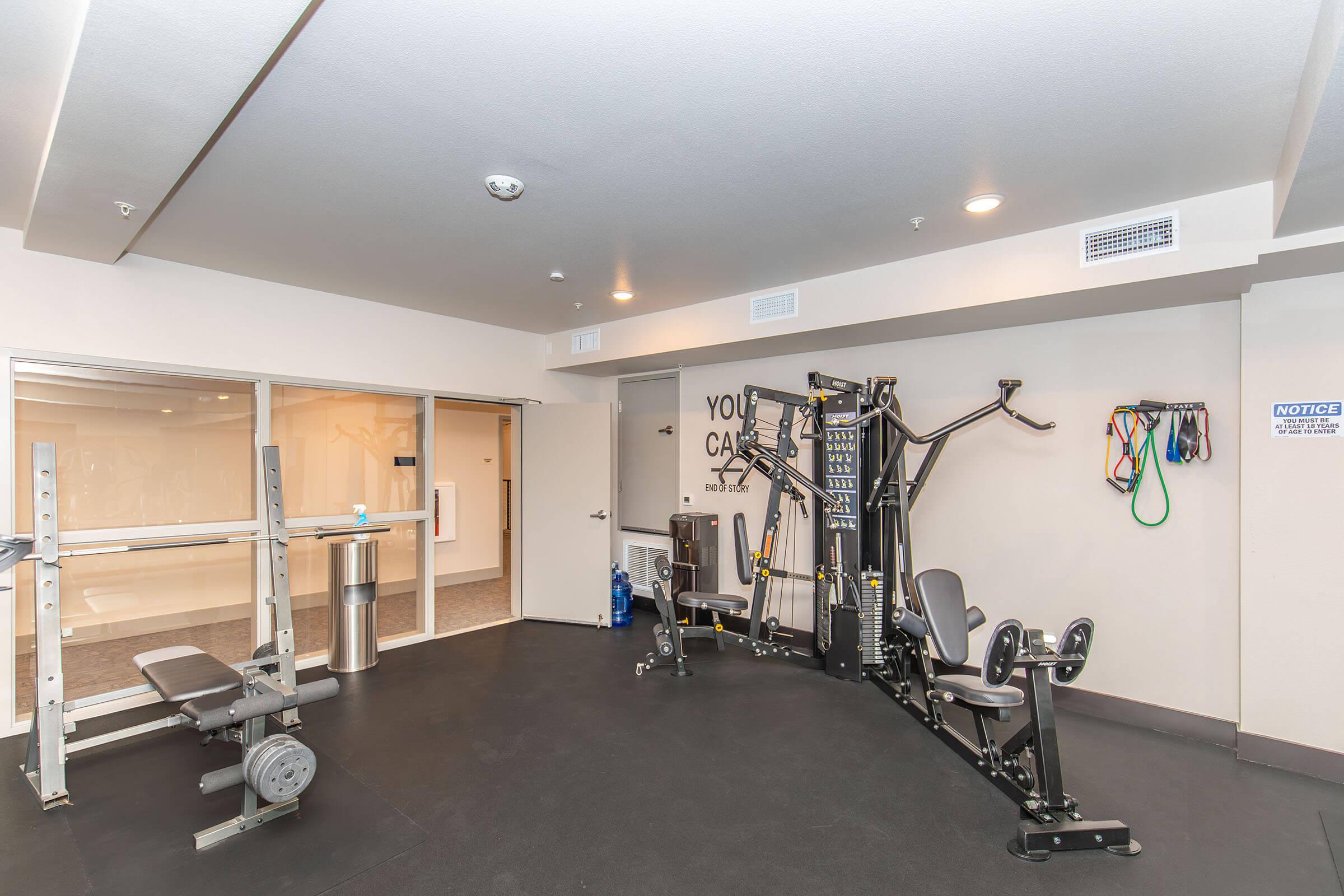A modern gym interior featuring strength training equipment. The space includes a multi-station gym machine, a weight bench, and resistance bands on the wall. Natural light enters from large windows, and the decor is minimalistic with an inspiring wall quote. Fitness accessories and a water dispenser are visible.