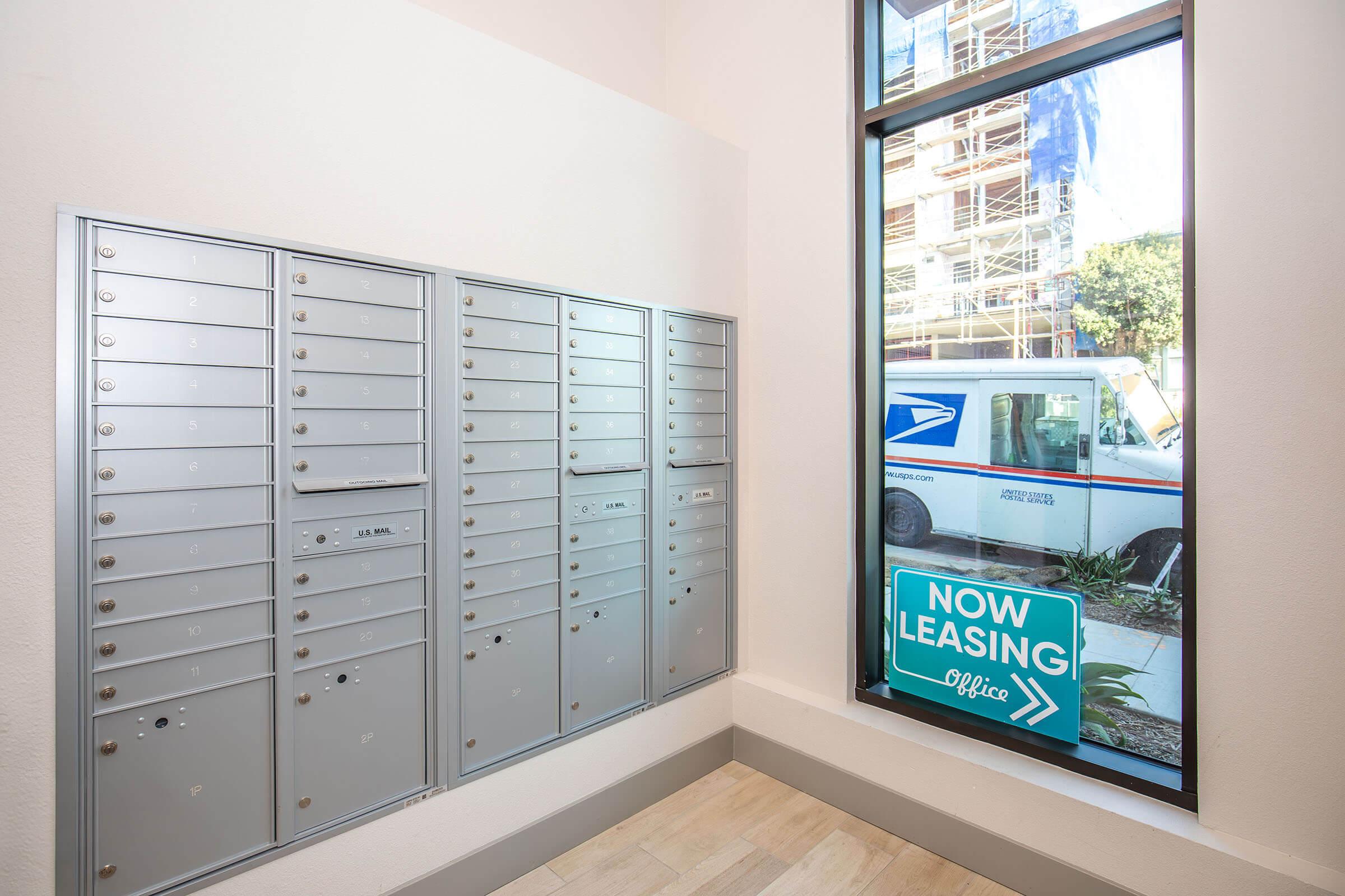 Interior view of a mailroom featuring a series of gray mailboxes on the left side. A large window on the right shows a USPS delivery truck outside, with construction scaffolding visible in the background. A blue sign reading "NOW LEASING Office" is placed on the window.