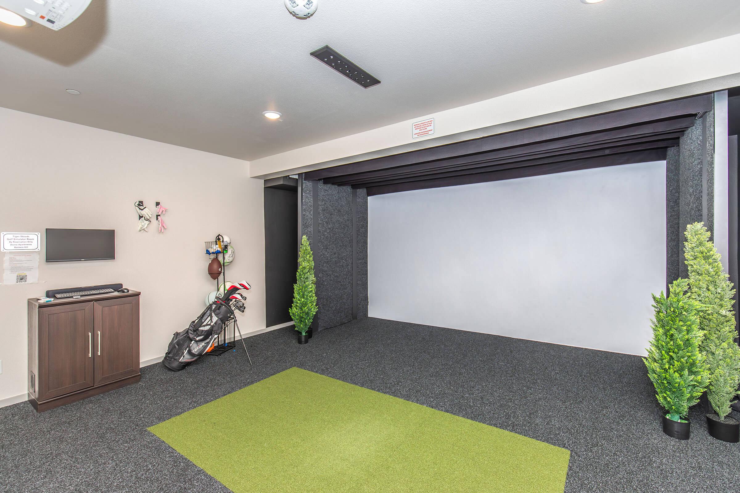 Indoor golf simulator room featuring a putting green, flat screen TV, golf clubs in a bag, and two artificial plants. The walls are lined with dark panels, and the space is designed for practicing golf swings and putting.