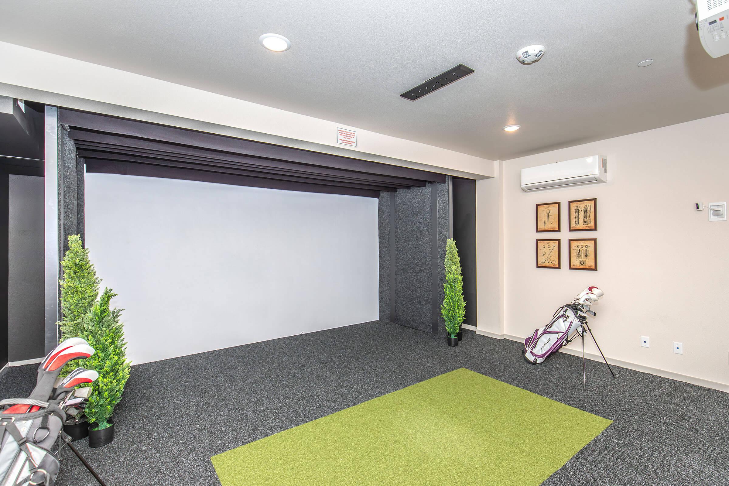 Indoor golf practice area featuring a putting green, golf clubs leaning against the wall, and a projector screen for virtual simulation. The room has dark walls with decorative art pieces, bright overhead lighting, and artificial plants positioned at the sides, creating a modern and inviting atmosphere.