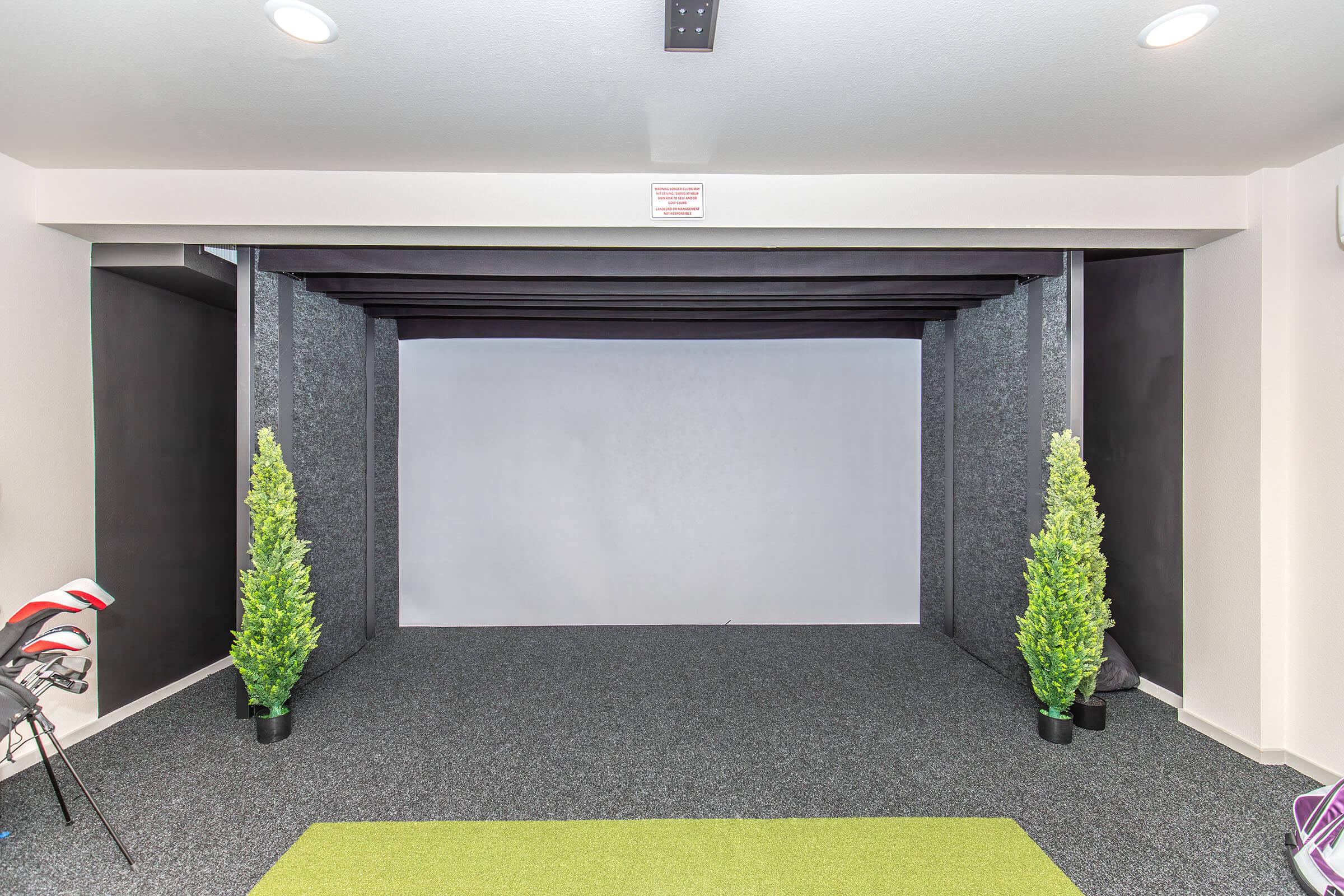 Indoor space featuring a textured gray wall, framed by dark beams. Two artificial green plants are placed in pots on either side. The floor is covered in dark carpet, and a green mat is laid out in the center. The area appears well-lit, creating a bright and inviting atmosphere.
