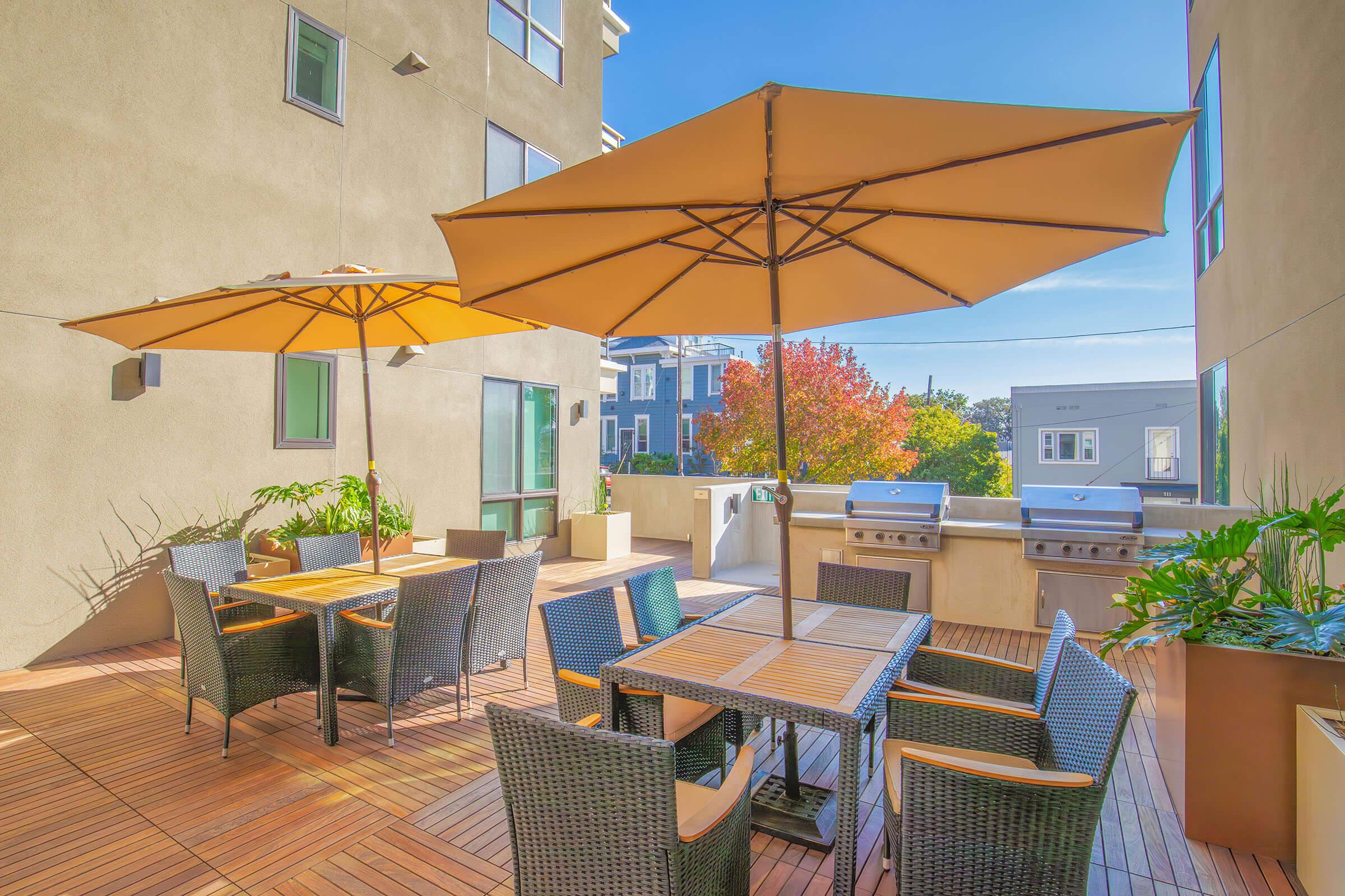 A modern outdoor patio with two large umbrellas providing shade over wicker tables and chairs. There are stainless steel grills in the background and planters with greenery, surrounded by a light-colored building with windows. The scene is bright and inviting, perfect for gatherings.