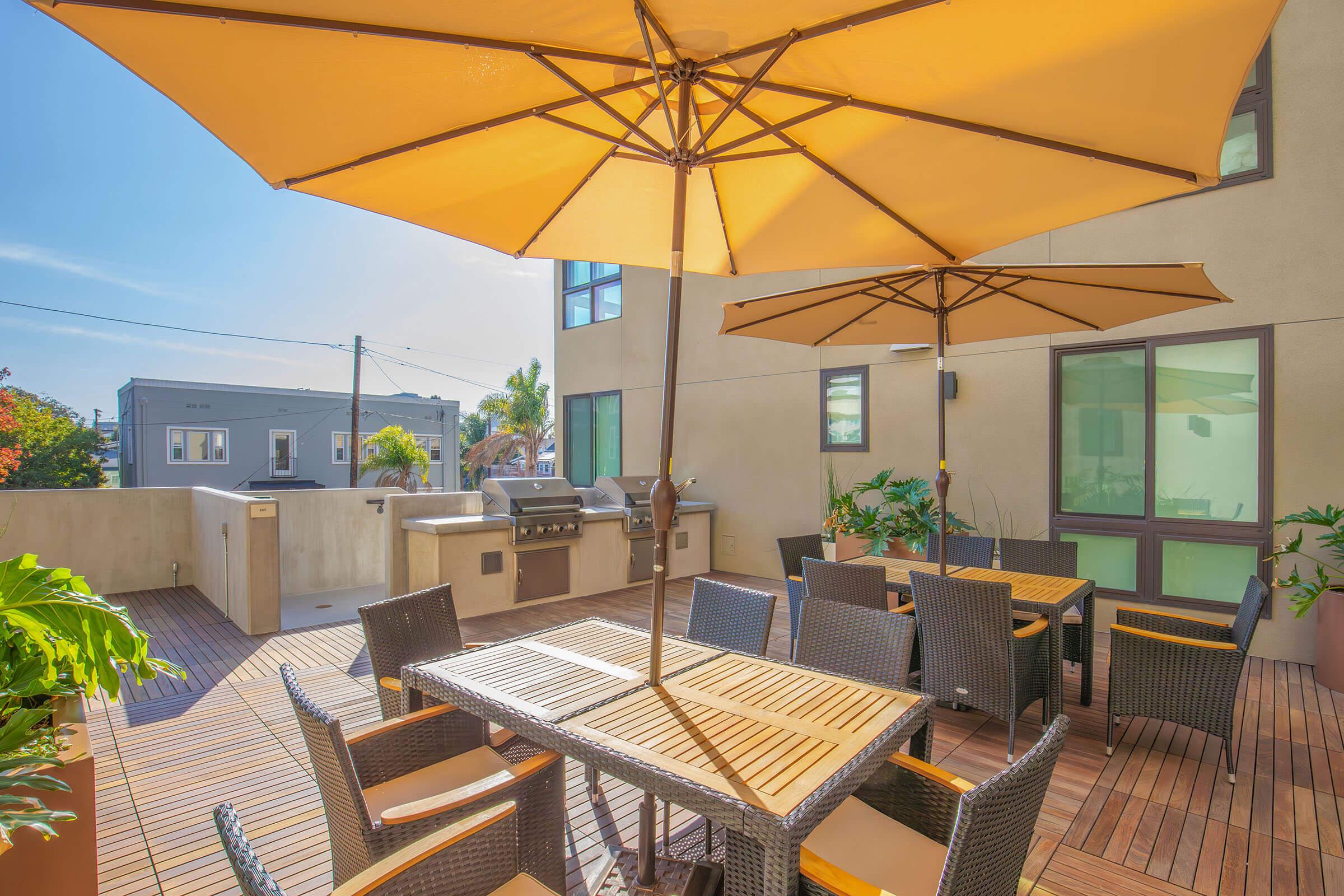 A sunny outdoor patio featuring a wooden dining table surrounded by wicker chairs. Large yellow umbrellas provide shade. In the background, there are green plants and a barbecue grill, with buildings and a clear blue sky visible.