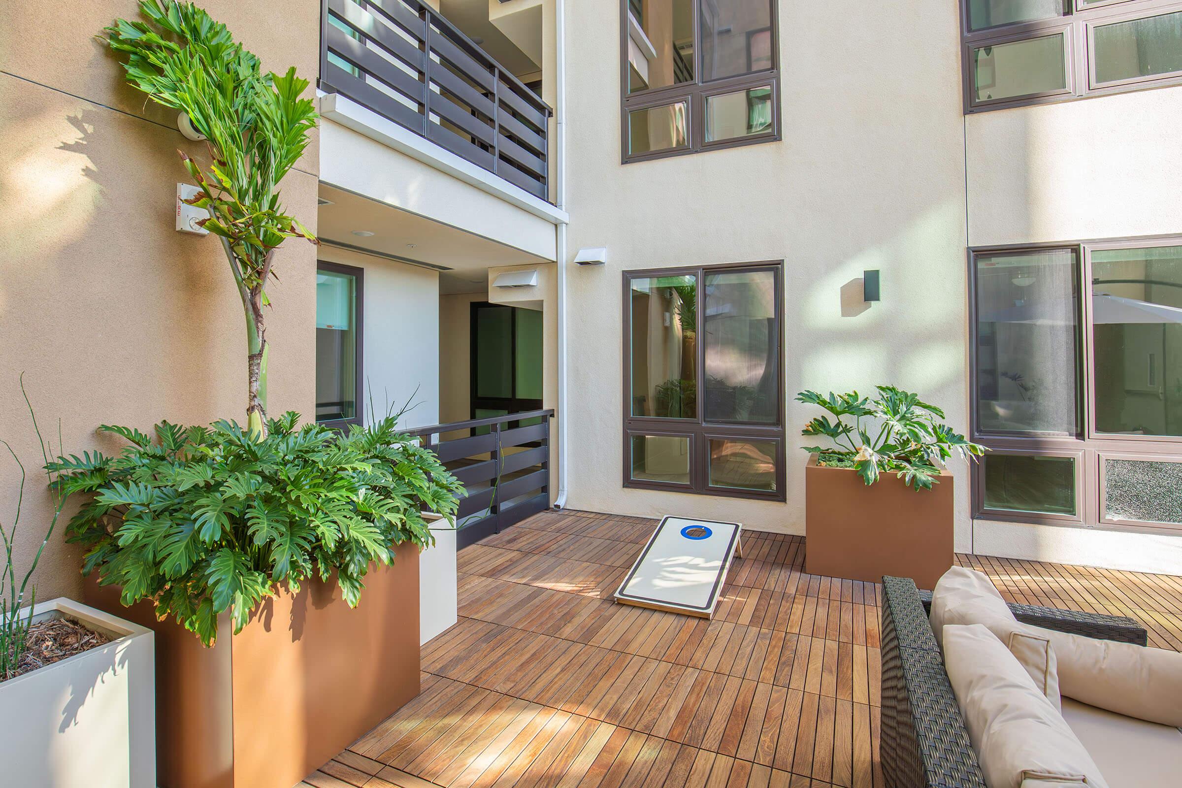 A modern courtyard with wooden flooring, featuring large planters with lush greenery. There are comfortable outdoor seating arrangements and a sleek, minimalist design. Sunlight streams in, highlighting the contemporary architecture and creating a bright, inviting atmosphere.