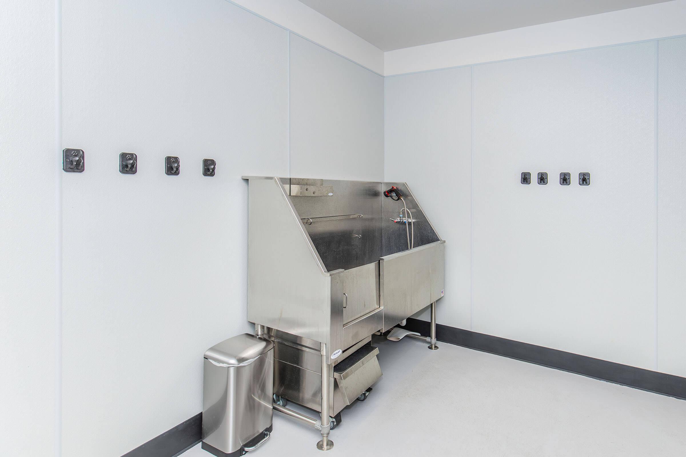 A clean, modern utility room featuring a stainless steel sink with a faucet, a trash can, and several electrical outlets on the pale blue walls. The floor is light-colored and the area appears well-lit and spacious, suitable for industrial or commercial use.