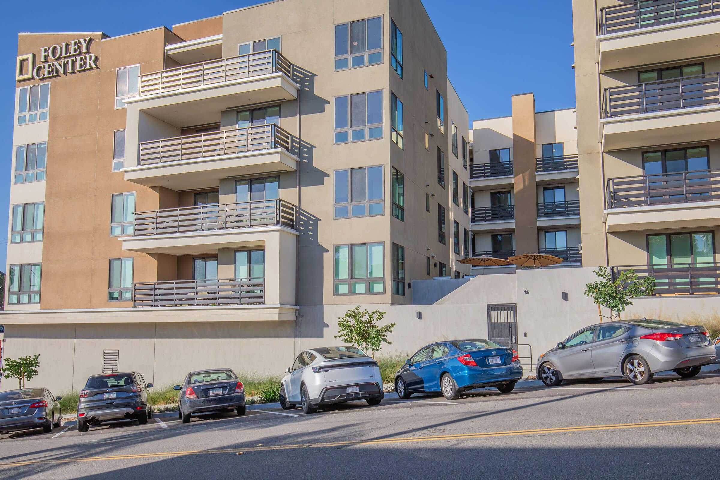 Residential building with the sign "Foley Center." The structure features multiple floors with balconies. Several cars are parked along the street in front of the building. The scene is set under a clear blue sky, highlighting the modern architecture and landscaping.