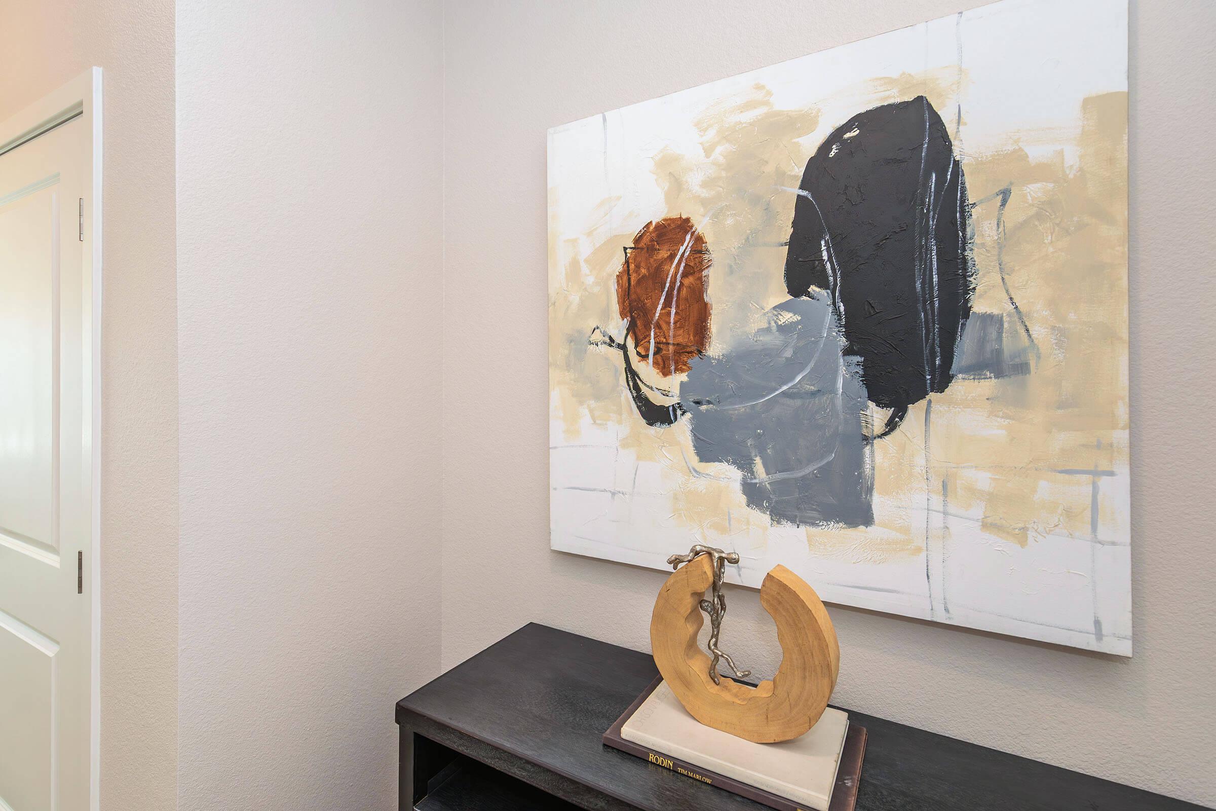 A modern interior scene featuring an abstract painting with neutral tones, showcasing bold black and orange shapes, hanging on a light-colored wall. Below, a wooden sculpture in a circular form rests on a dark console table, enhancing the contemporary aesthetic of the space.