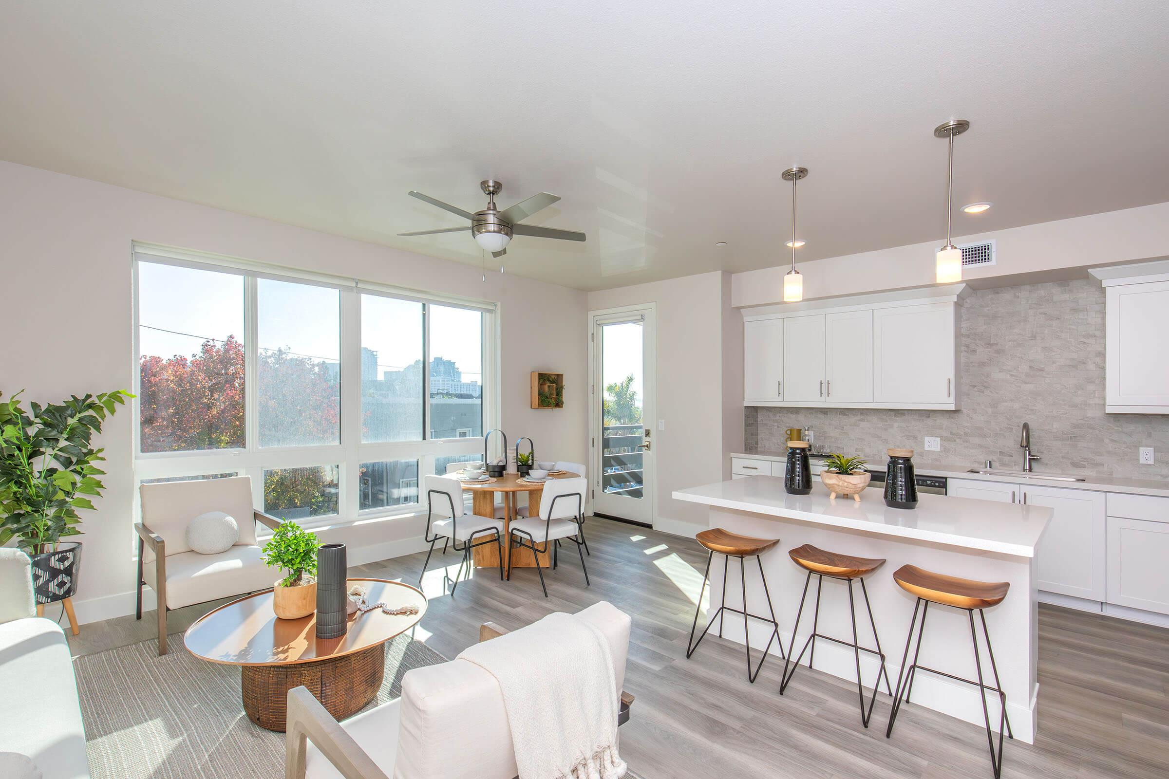 A modern, bright apartment interior featuring an open kitchen and living area. The kitchen has white cabinets and bar seating with three stools. A dining table is set for four, and large windows allow natural light in, showcasing a vase of plants and minimalistic decor.