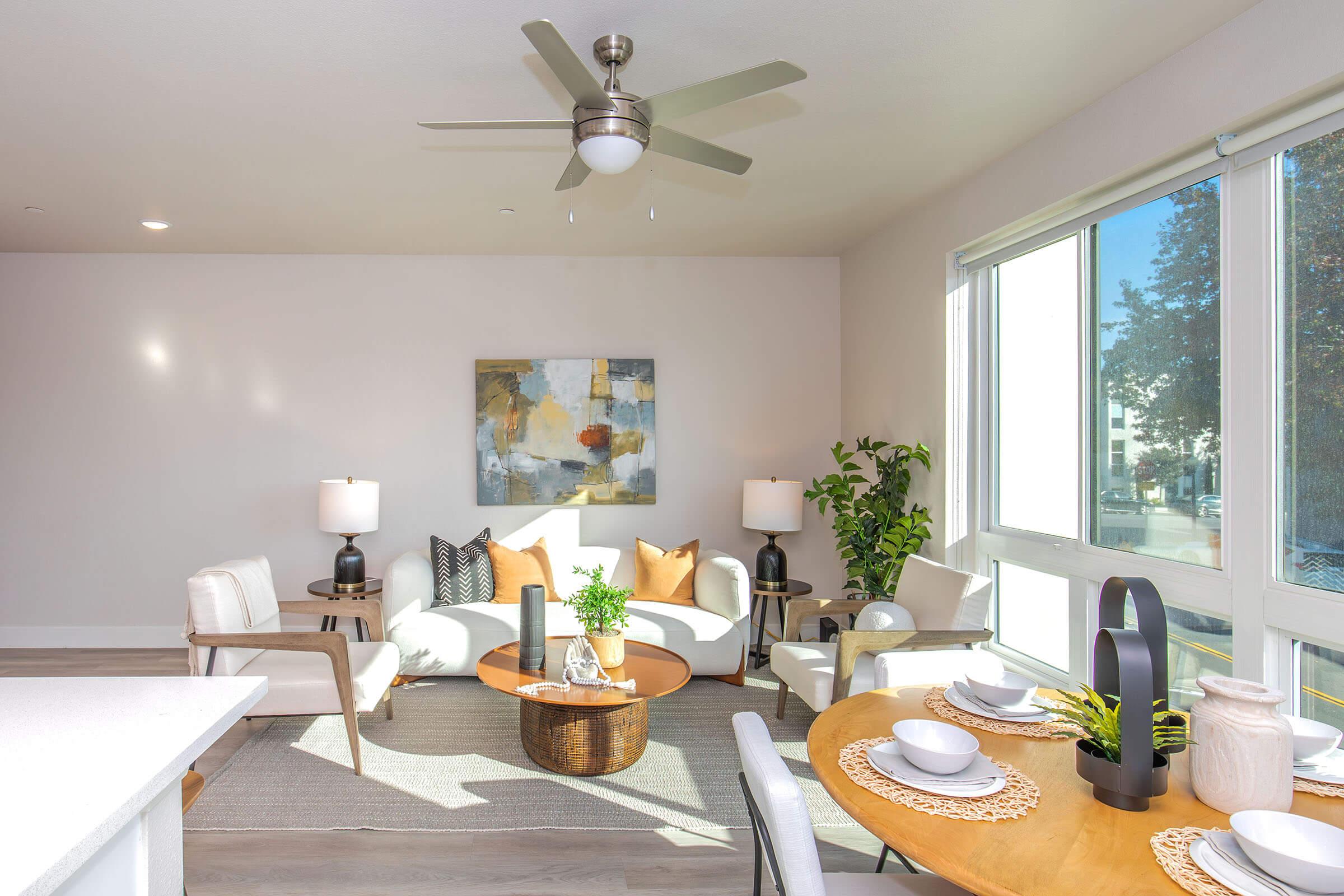 A bright and modern living space featuring a comfortable seating area with white chairs and orange accent pillows, a round wooden coffee table, a dining table set for two, and large windows letting in natural light. Decorative plants and art add a cozy touch to the room.