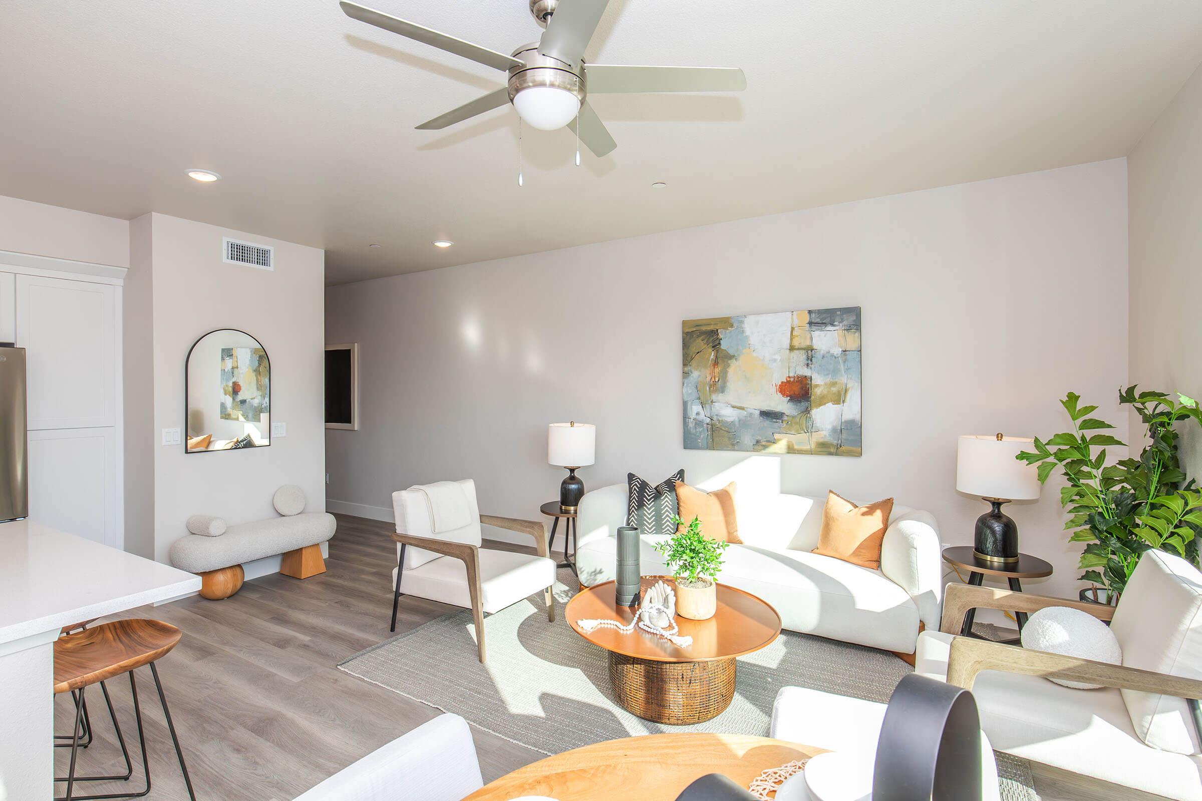 Bright living room featuring modern furniture, including white sofas and a circular coffee table. Natural light fills the space, highlighting a colorful abstract painting on the wall. The room includes a fan, stylish lamps, and a decorative plant, creating a cozy and inviting atmosphere.