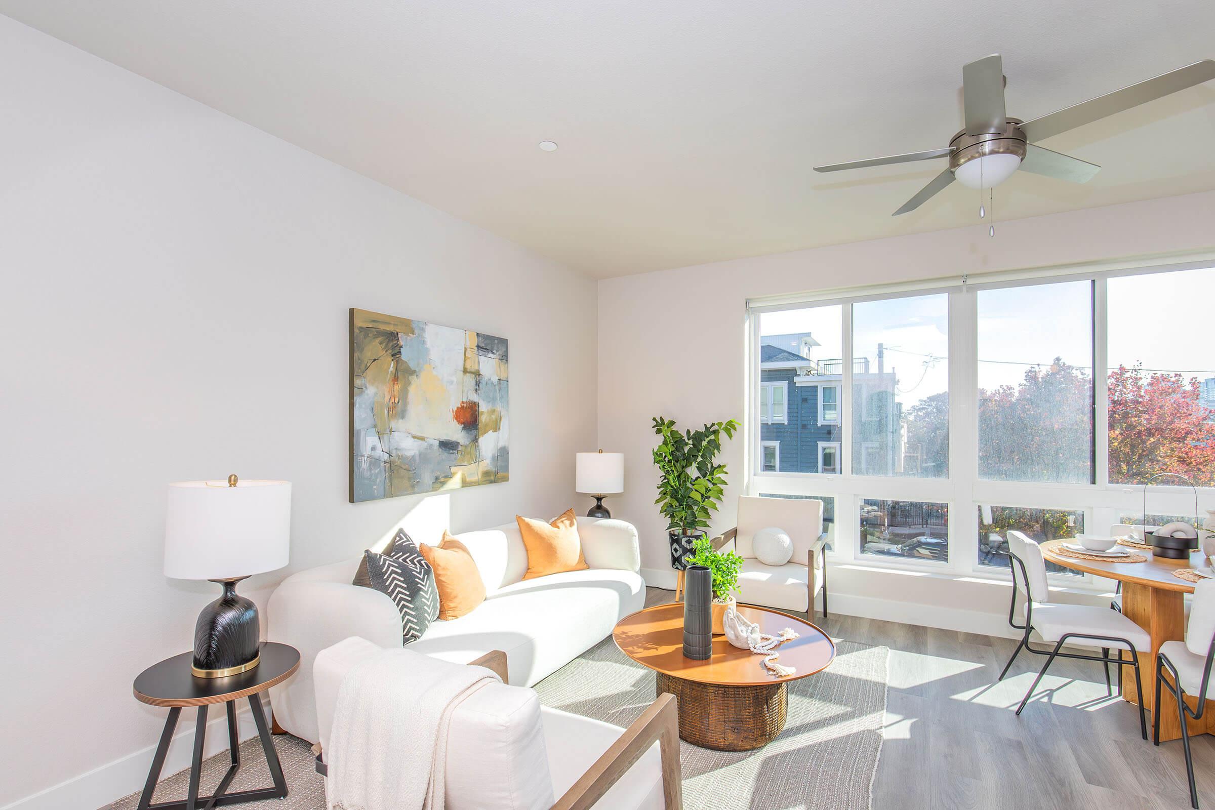 A bright and modern living room featuring a white sofa with orange and black accents, a round wooden coffee table, a potted plant, and a ceiling fan. Natural light flows in through large windows, illuminating a dining area with a wooden table and stylish decor.