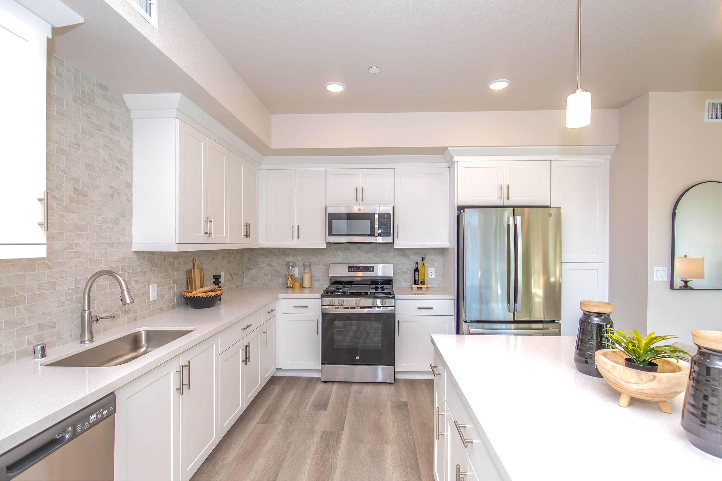 Modern kitchen featuring white cabinetry, stainless steel appliances, and a stone backsplash. The space includes a double-basin sink, a gas stove with an oven, and a refrigerator. Light wood flooring complements the bright, airy atmosphere, enhanced by natural light from a window. Decorative items are placed on the countertop.