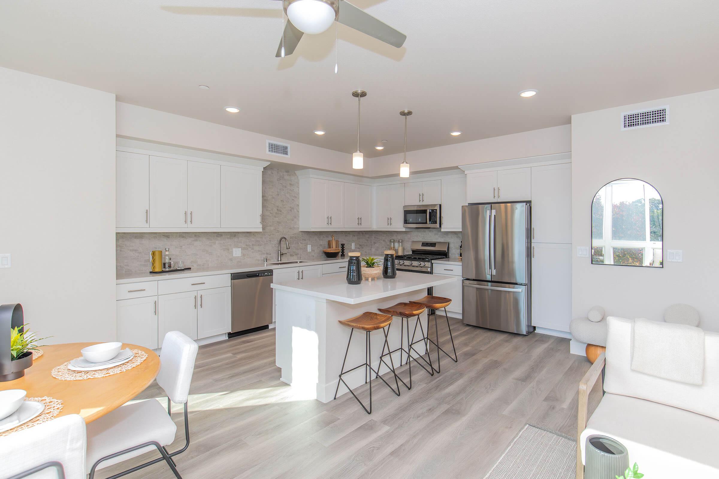 Modern kitchen in an open-concept living space, featuring white cabinetry, stainless steel appliances, and a large island with seating. Natural light floods the room through large windows, complementing the neutral color palette and contemporary decor. Dining table set with plates and a centerpiece enhances the inviting atmosphere.