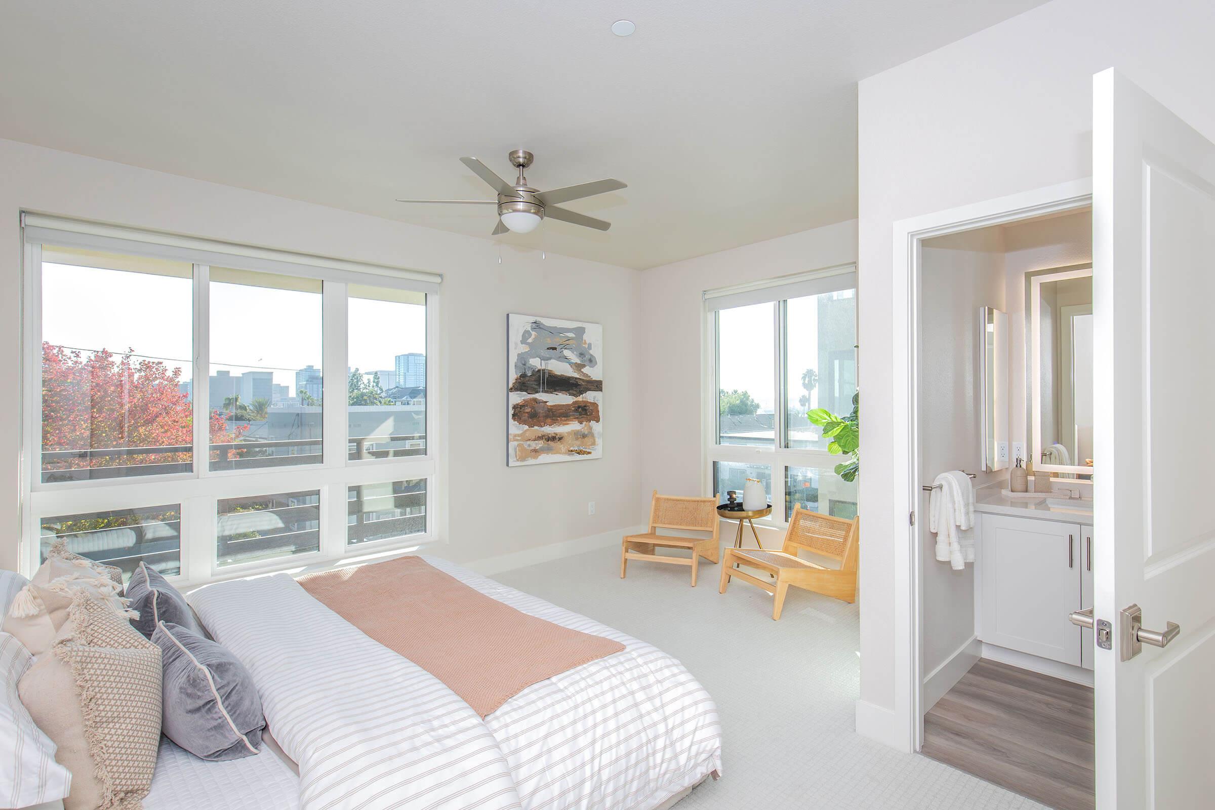 Bright and modern bedroom featuring a large bed with decorative pillows, a ceiling fan, and two wooden chairs near a window. Natural light fills the space, which includes an en-suite bathroom visible through an open door. The decor is minimalist with a touch of nature visible outside.