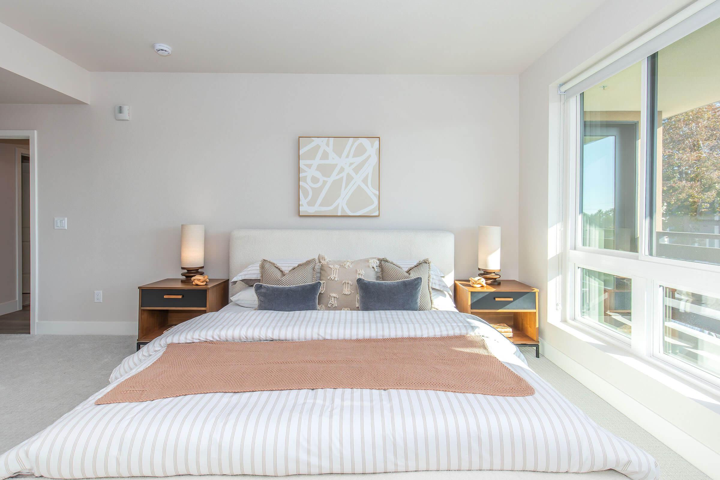A modern bedroom featuring a large bed with a striped duvet, decorative pillows, and a cozy throw. There are matching nightstands with lamps on either side of the bed. A piece of abstract art hangs above the headboard, and large windows offer natural light and a view of the outdoors.