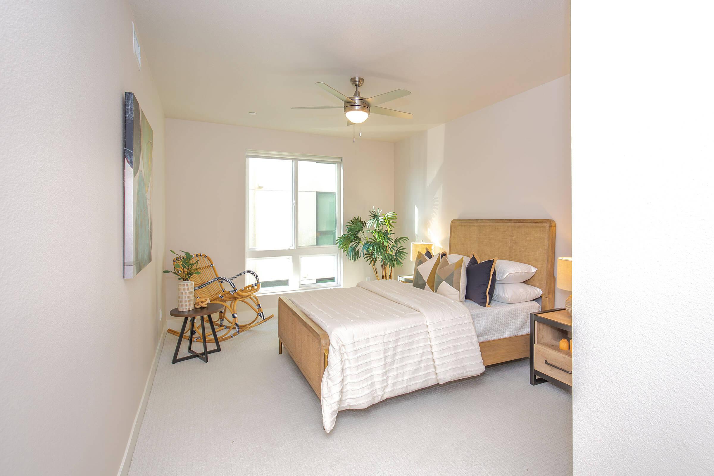 A neatly arranged bedroom featuring a comfortable queen-sized bed with white linens, decorative pillows, a bedside table with a lamp, and a modern ceiling fan. Natural light streams in through a large window, complemented by a small plant and a cozy reading chair in the corner.