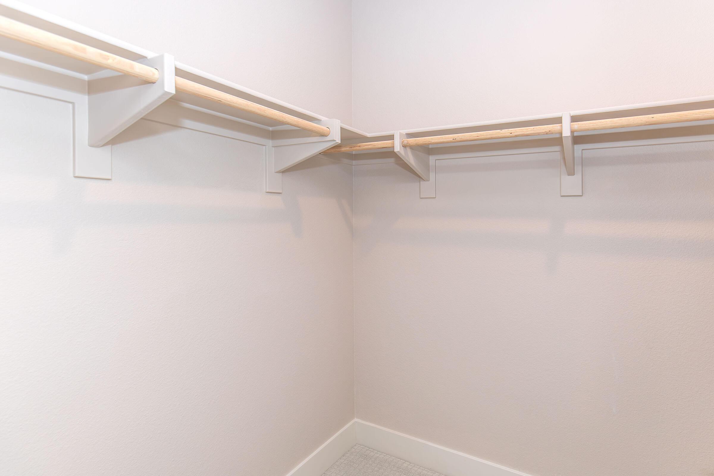 Empty walk-in closet featuring two wooden hanging rods supported by white brackets, set in a plain, light-colored wall. The floor is tiled with a light pattern, and the space is well-lit, creating a clean and minimalistic look. Ideal for organizing clothing and accessories.