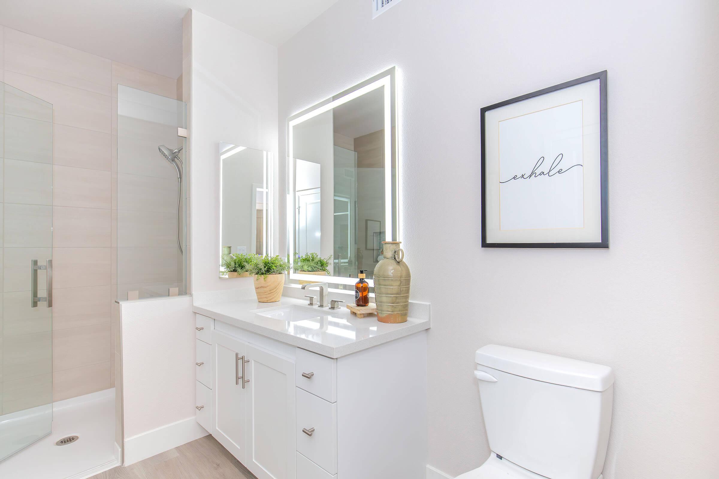 A modern bathroom featuring a white vanity with dual mirrors and stylish lighting, a potted plant, decorative bottle, and a framed print with the word "exhale." The space includes a shower area with glass doors and neutral-colored walls and flooring, creating a serene and clean aesthetic.