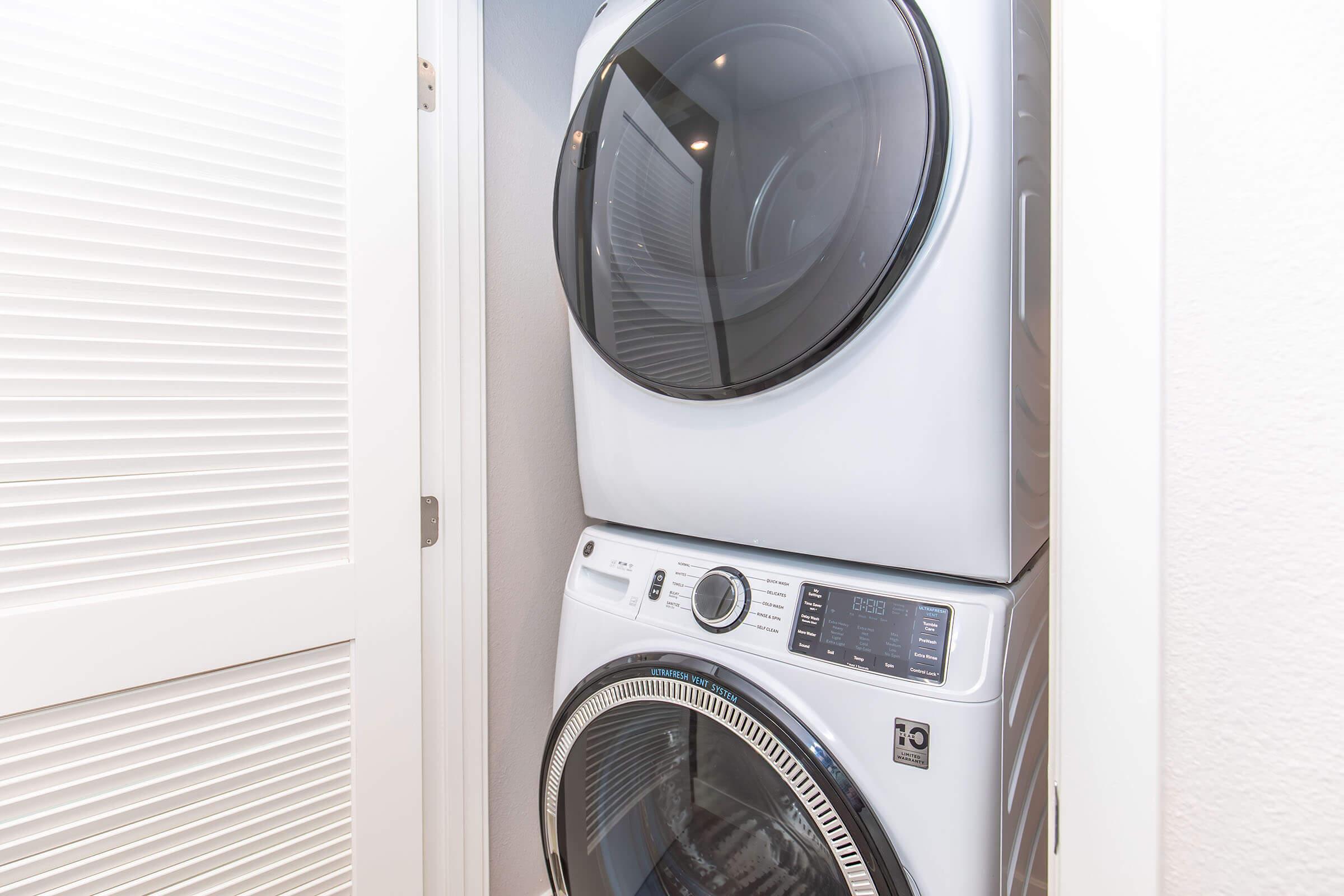 A stacked washer and dryer set in a small laundry nook, featuring a modern design with a white exterior. The washer is at the bottom and the dryer on top, both with glass doors. The space has sliding doors for easy access and organization.