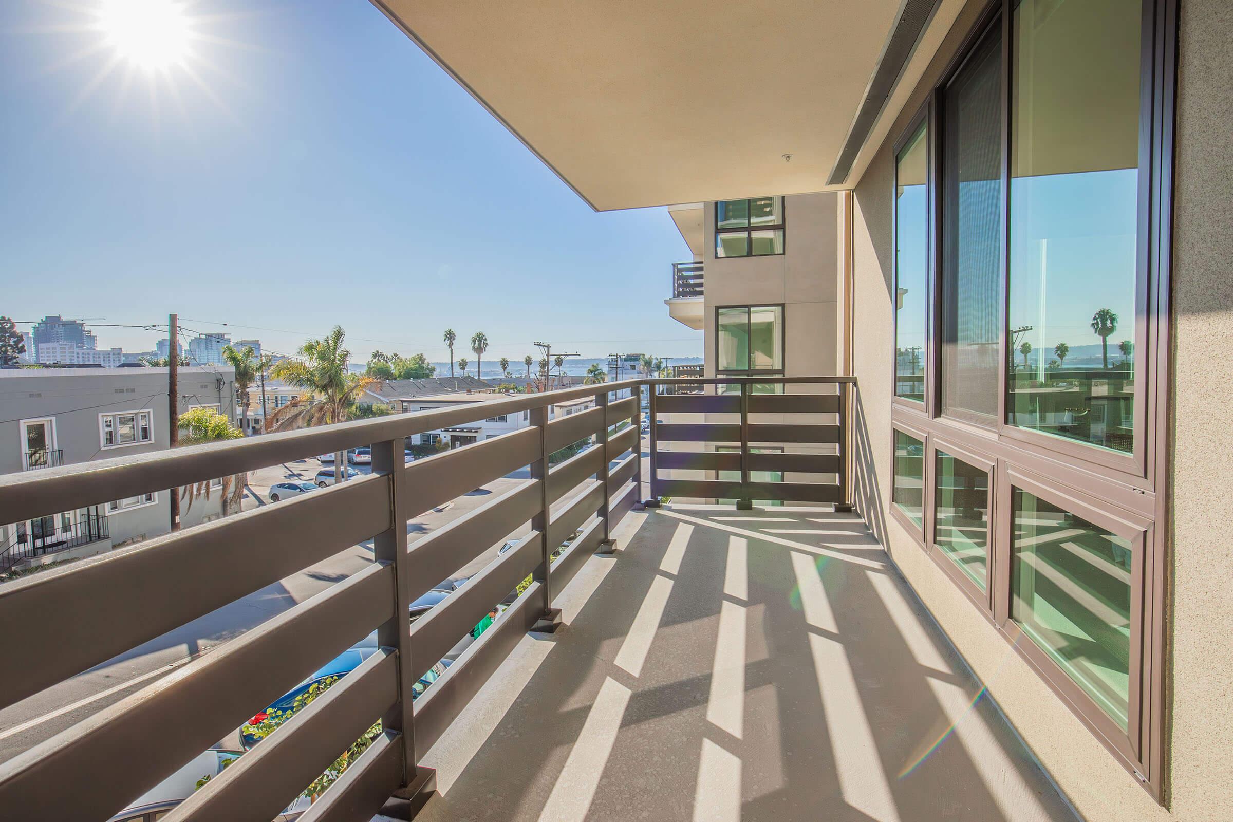 A sunny balcony view with a railing, overlooking a street lined with palm trees. The sunlight casts shadows on the floor, and in the distance, buildings and a clear blue sky are visible, creating a vibrant and inviting atmosphere.