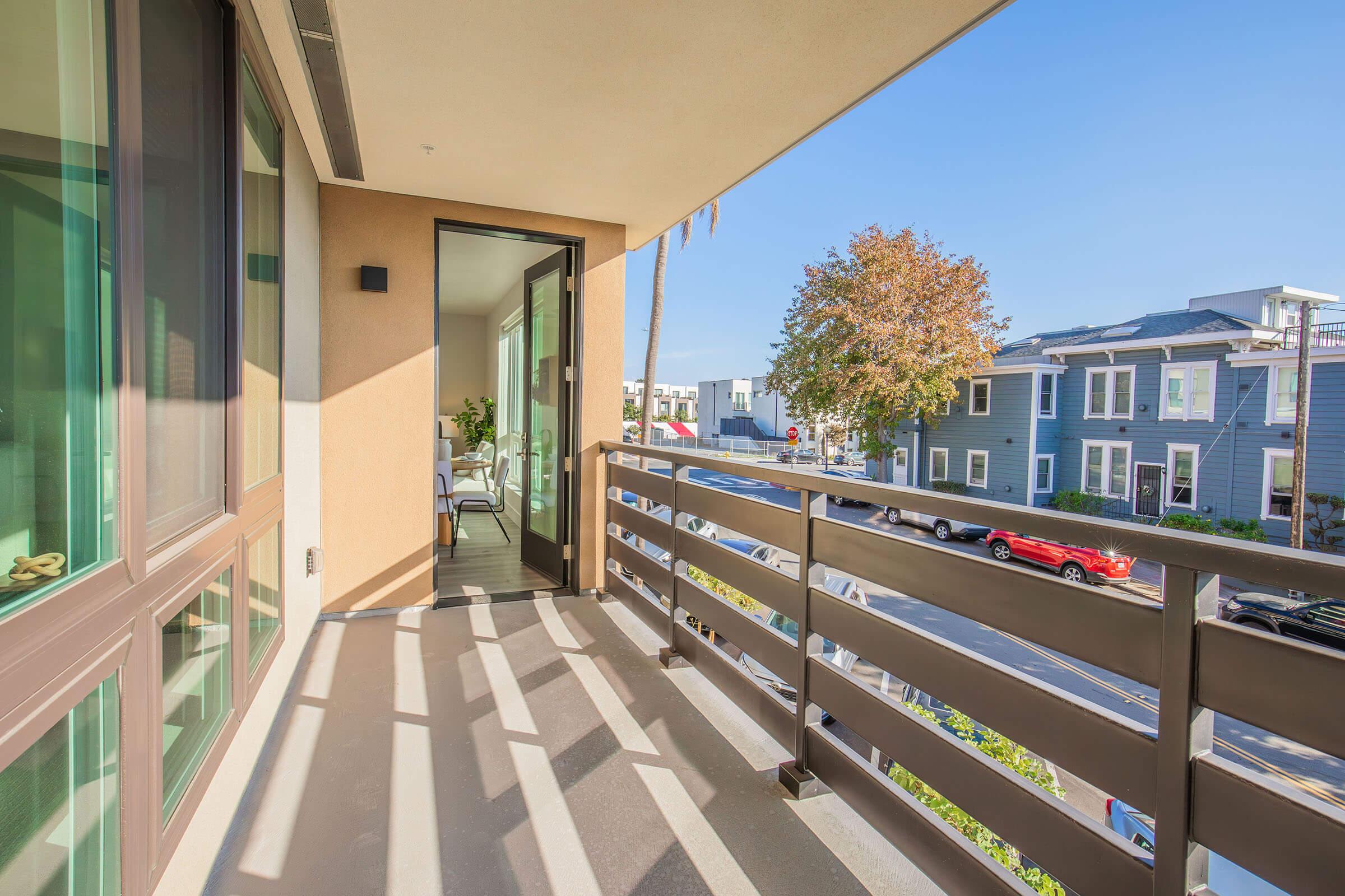 A modern balcony with a view of a tree and residential buildings. The space features a sleek railing and shadows cast by sunlight. Inside, there is a glimpse of a bright, inviting interior with furniture.