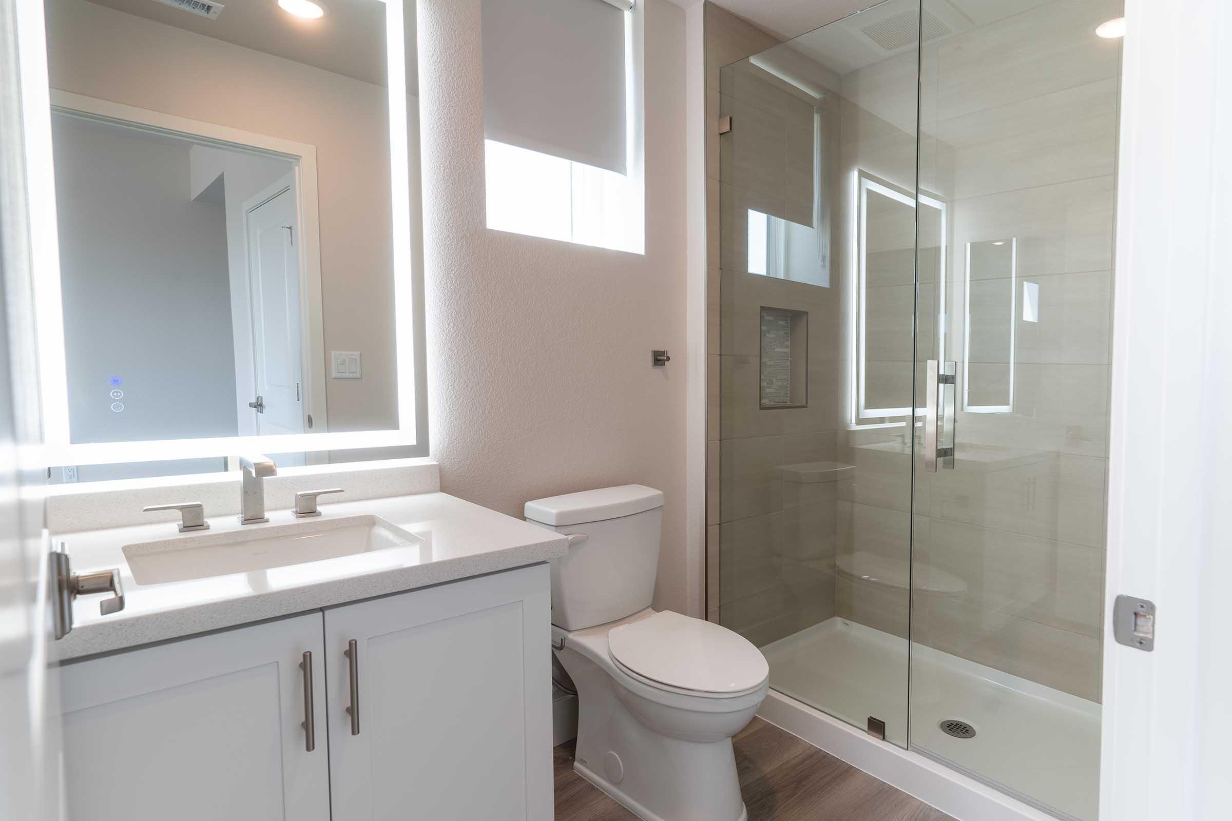 A modern bathroom featuring a glass shower, a sleek vanity with a double sink, a white toilet, and neutral-toned walls. The space is well-lit with LED-backlit mirrors and has a clean, contemporary design.