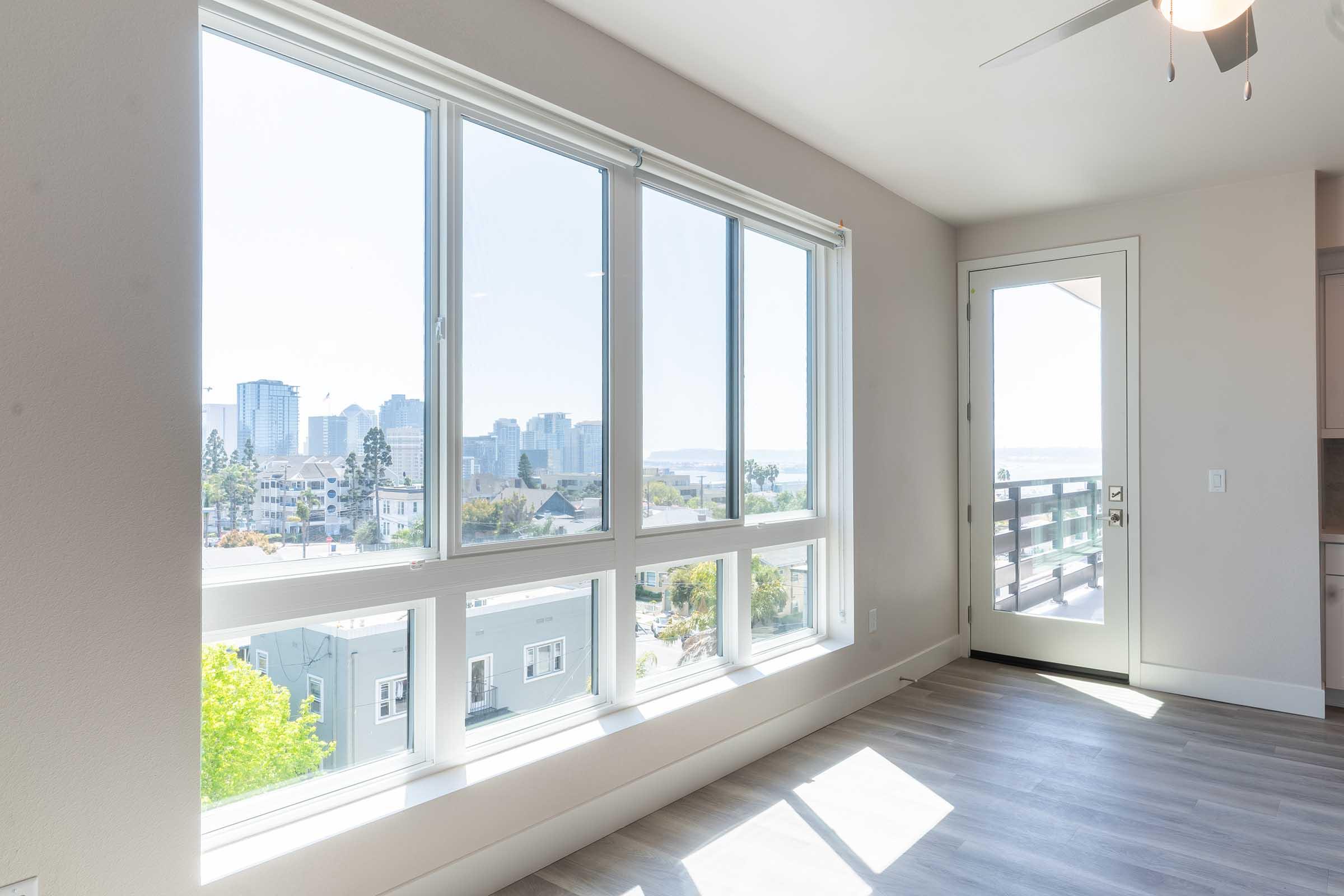 Bright, modern interior with large windows showcasing a city skyline view. The open space features light-colored walls and flooring, allowing natural light to fill the room. A door opens to an outdoor area, enhancing the airy ambiance. Urban landscape visible beyond the windows.