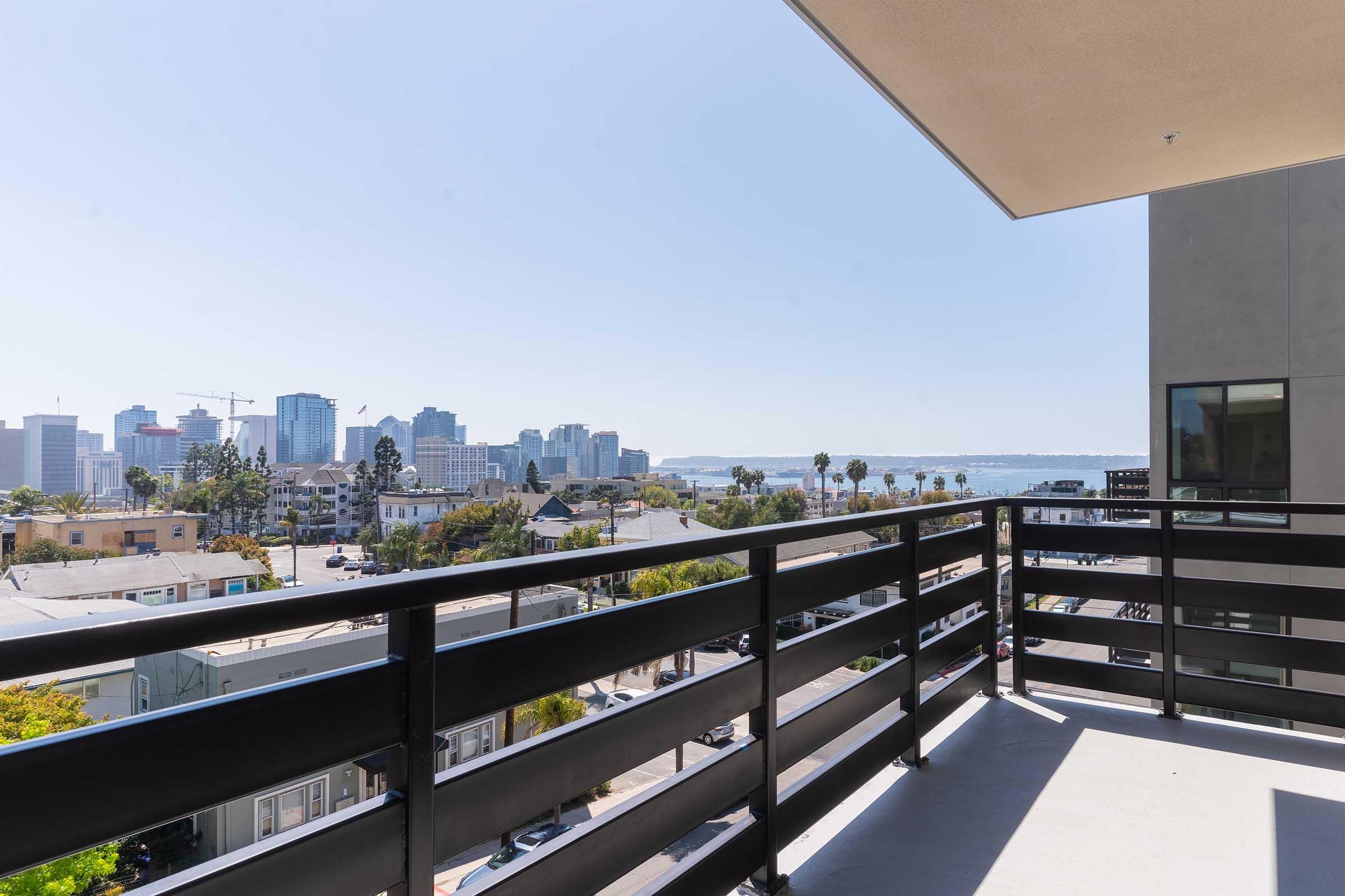 View from a balcony showcasing a city skyline with tall buildings, palm trees, and waterfront in the background under a clear blue sky. The foreground features a modern railing, emphasizing the urban landscape and serene coastal setting.