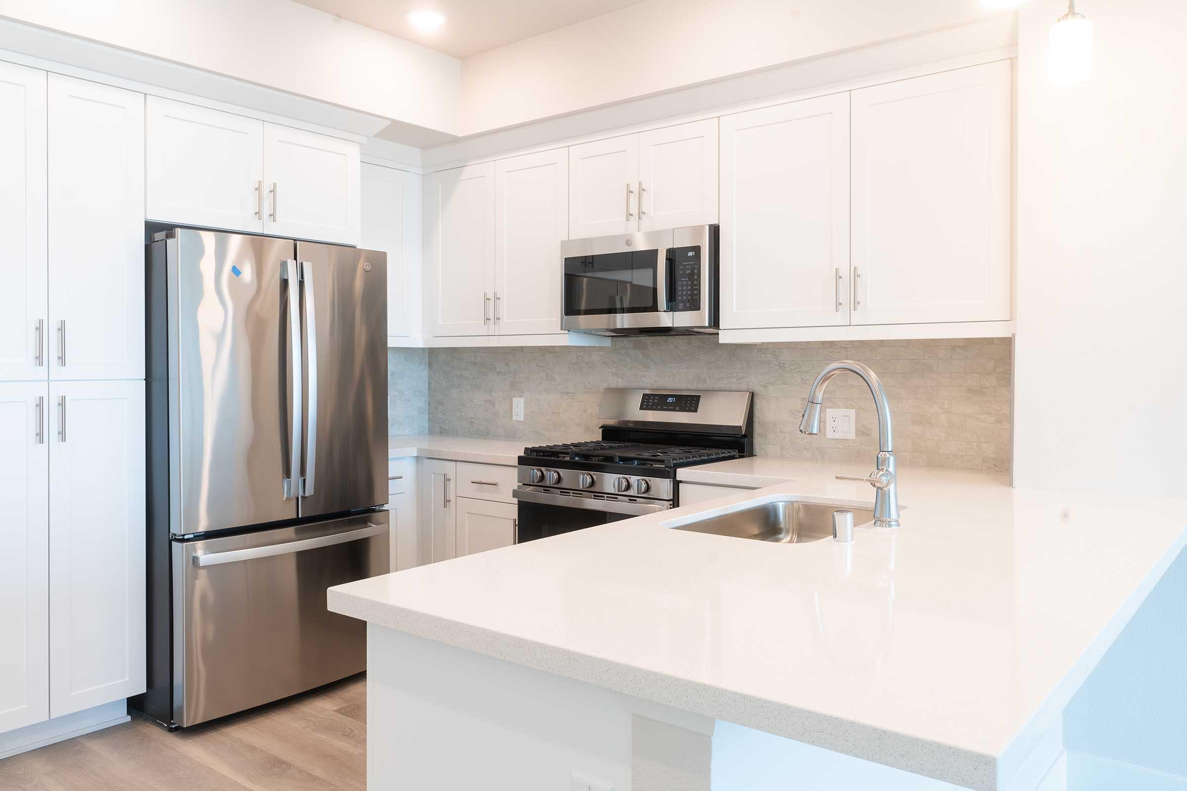 A modern kitchen featuring white cabinetry, stainless steel appliances including a refrigerator and microwave, a gas stovetop, and a sink on a light-colored countertop. The design is bright and airy with natural light illuminating the space.