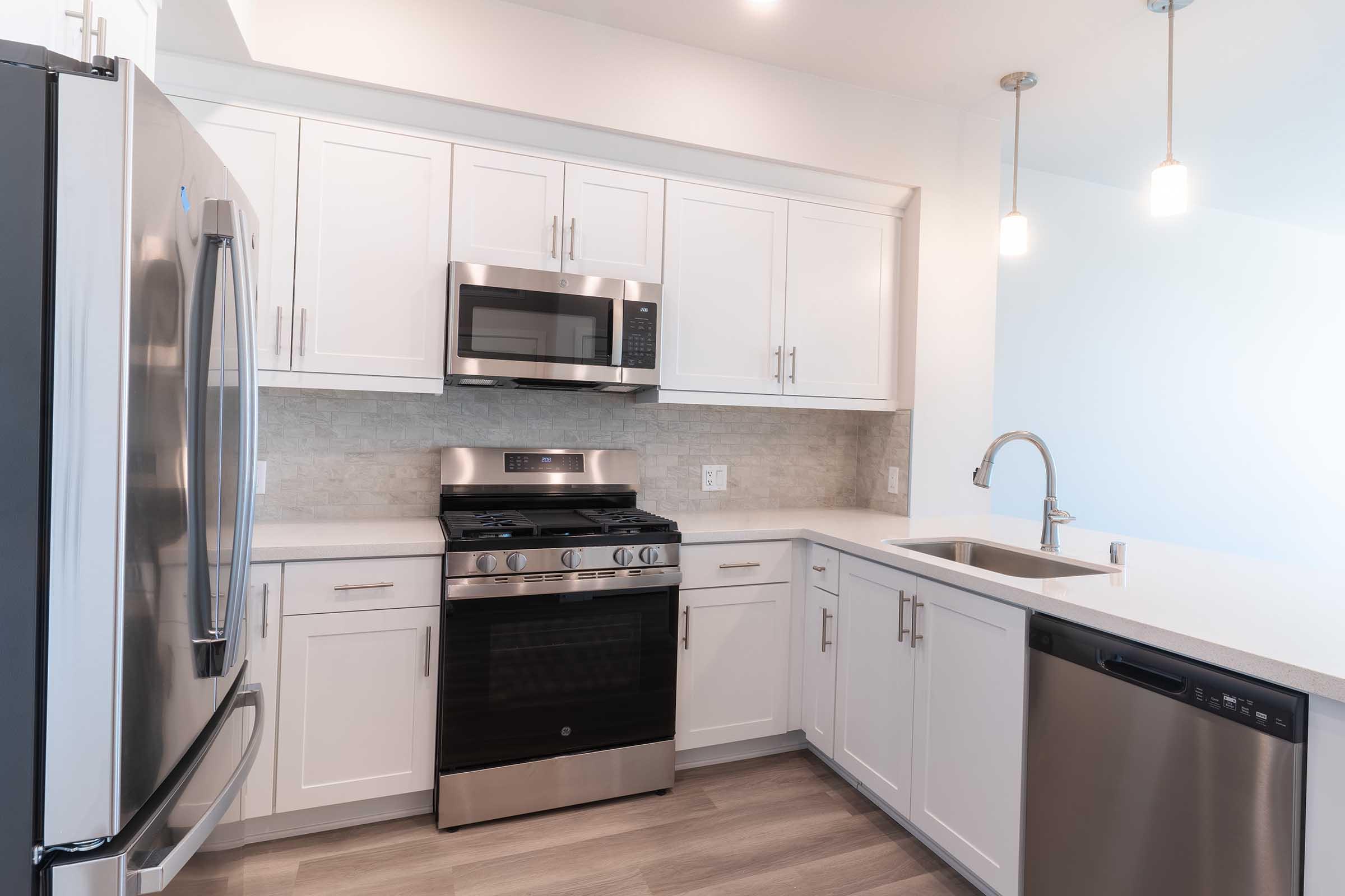 Modern kitchen featuring stainless steel appliances, including a fridge, oven, and microwave. The cabinetry is white with sleek handles, complemented by a light countertop. A stainless steel sink is under a stylish faucet, and there are two pendant lights hanging above the counter.