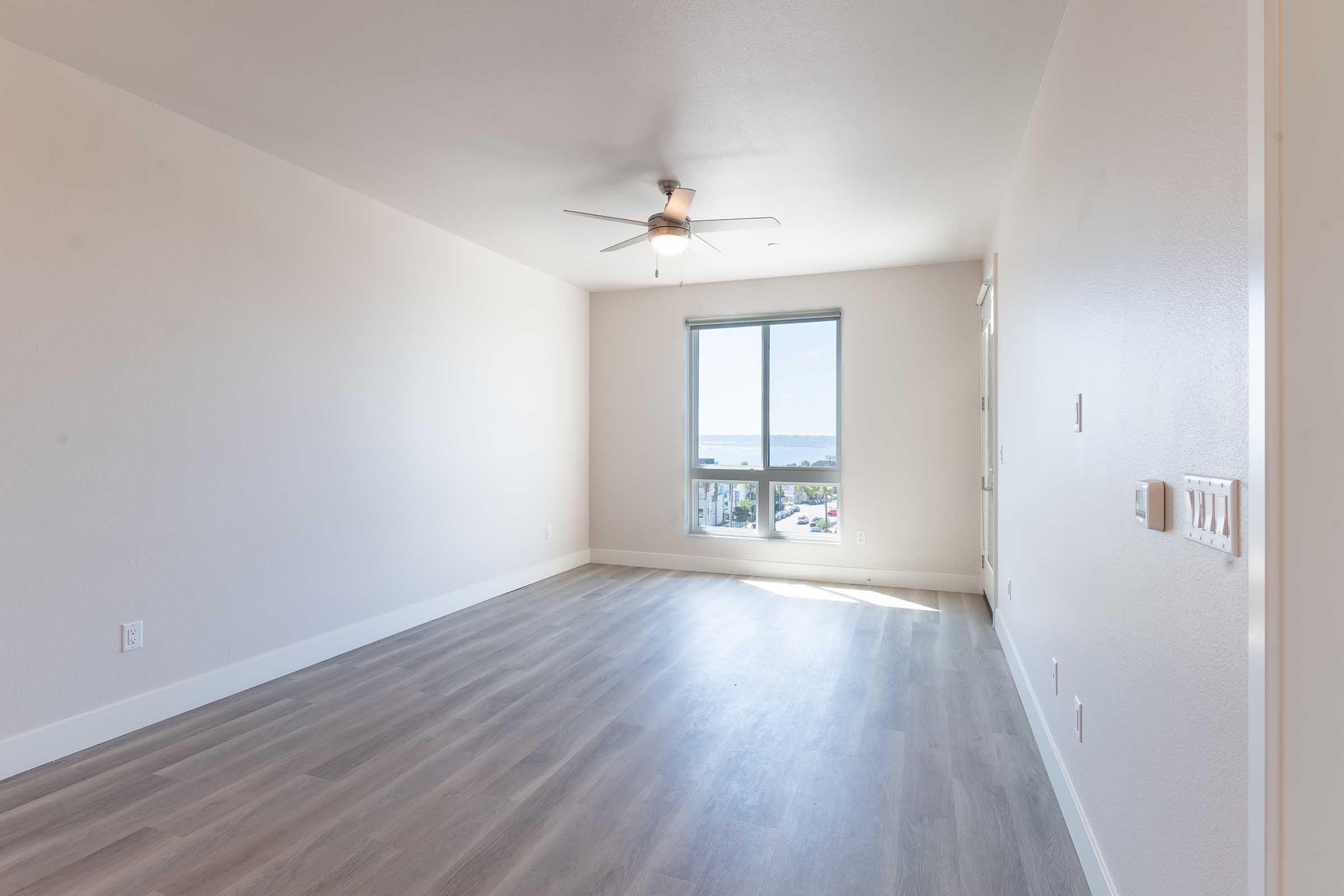A spacious, bright living room with light-colored walls and a ceiling fan. The floor is finished with light wood-like flooring. A large window allows natural light to enter, offering a view of the outdoors, while a door leads to another room.