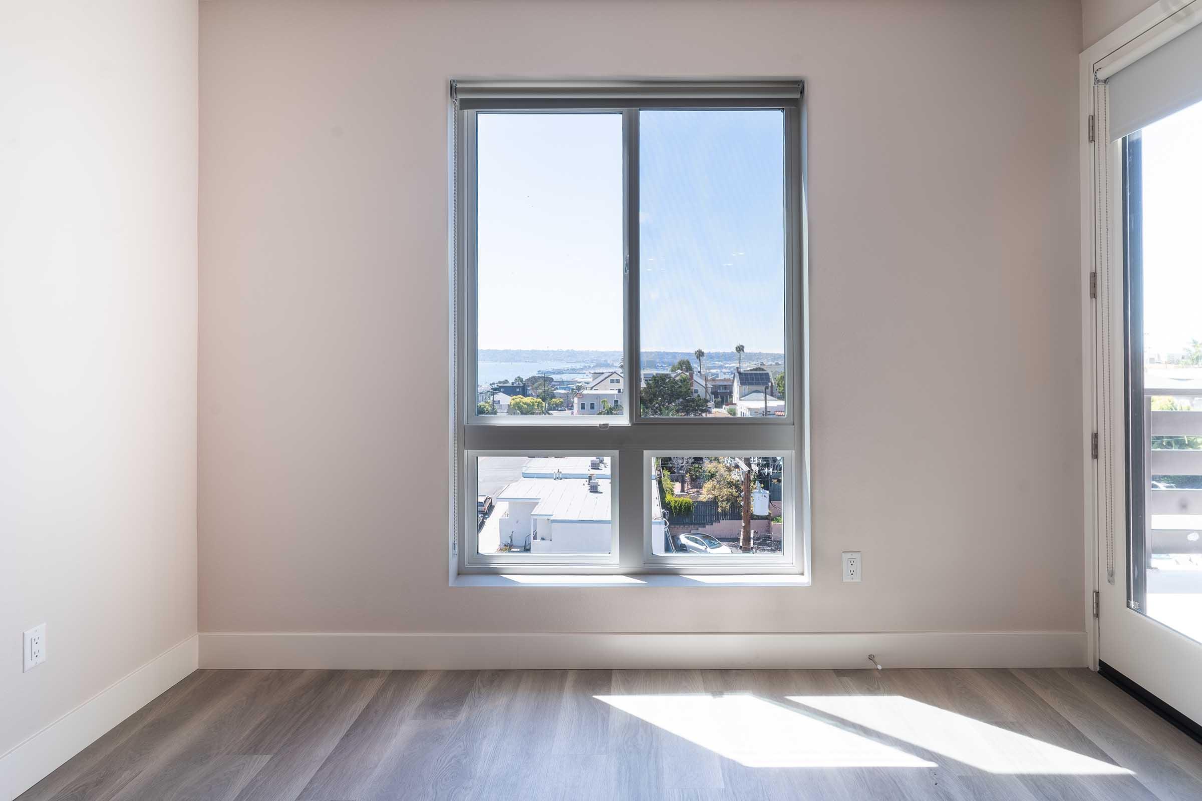 A bright, empty room with light-colored walls and a large window showcasing a view of nearby buildings and a clear sky. The floor is made of light wood, and there is a sliding door leading to a balcony or outdoor area. The overall atmosphere is spacious and inviting.
