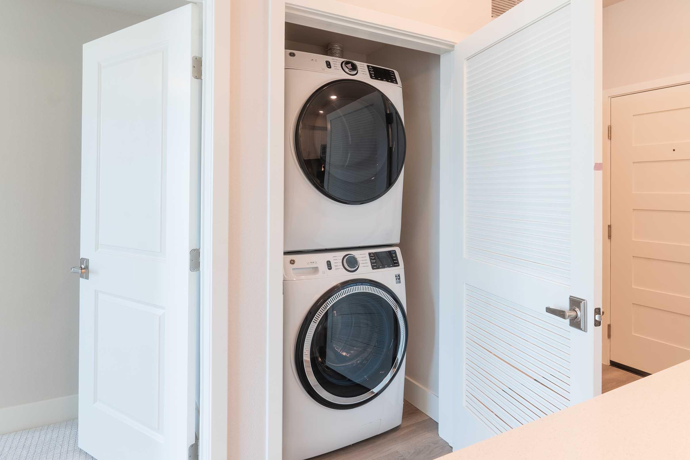 A compact laundry area featuring a stacked washer and dryer unit built into a closet space, with white doors partially open. The walls are painted in a light color, and the flooring is a light wood. The doors have modern hardware, giving a clean and efficient look to the laundry setup.