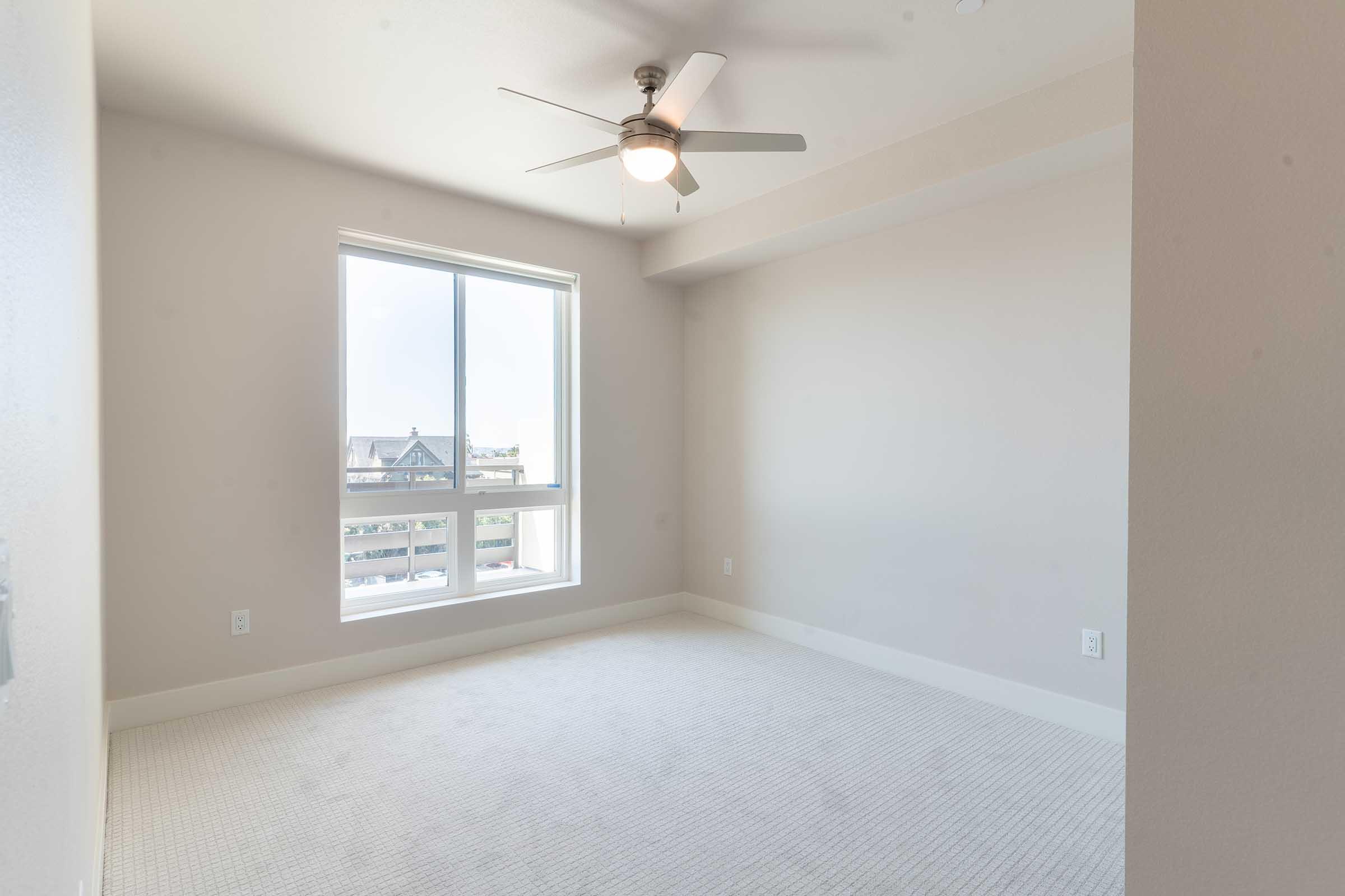 A spacious, empty room featuring light-colored walls and plush carpet. It has a ceiling fan and a large window providing natural light and a view of the outdoors. The room is well-lit and has a modern, minimalistic design, making it suitable for various uses.