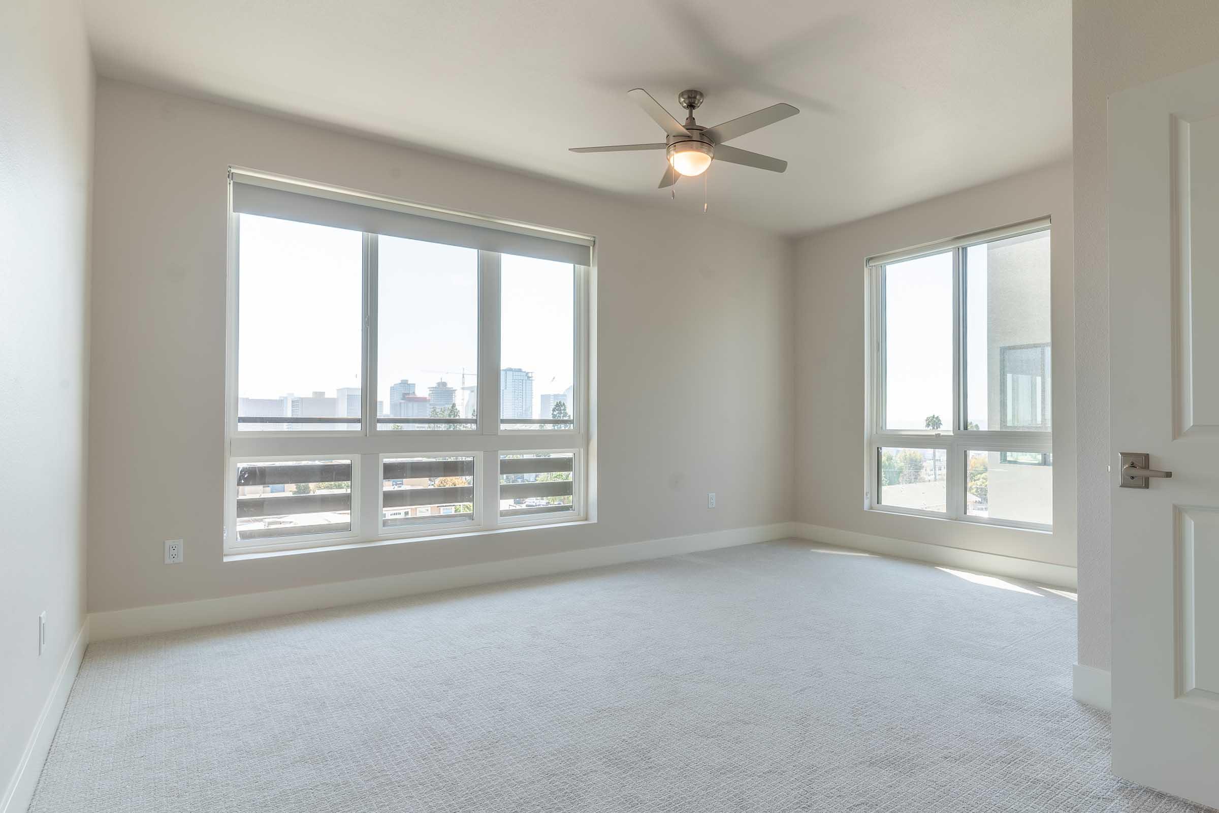 A bright, empty room with large windows allowing natural light. The walls are painted a light color, and the floor is covered with carpet. A ceiling fan is installed overhead, and there’s a view of a city skyline through the windows. The overall atmosphere is spacious and airy.