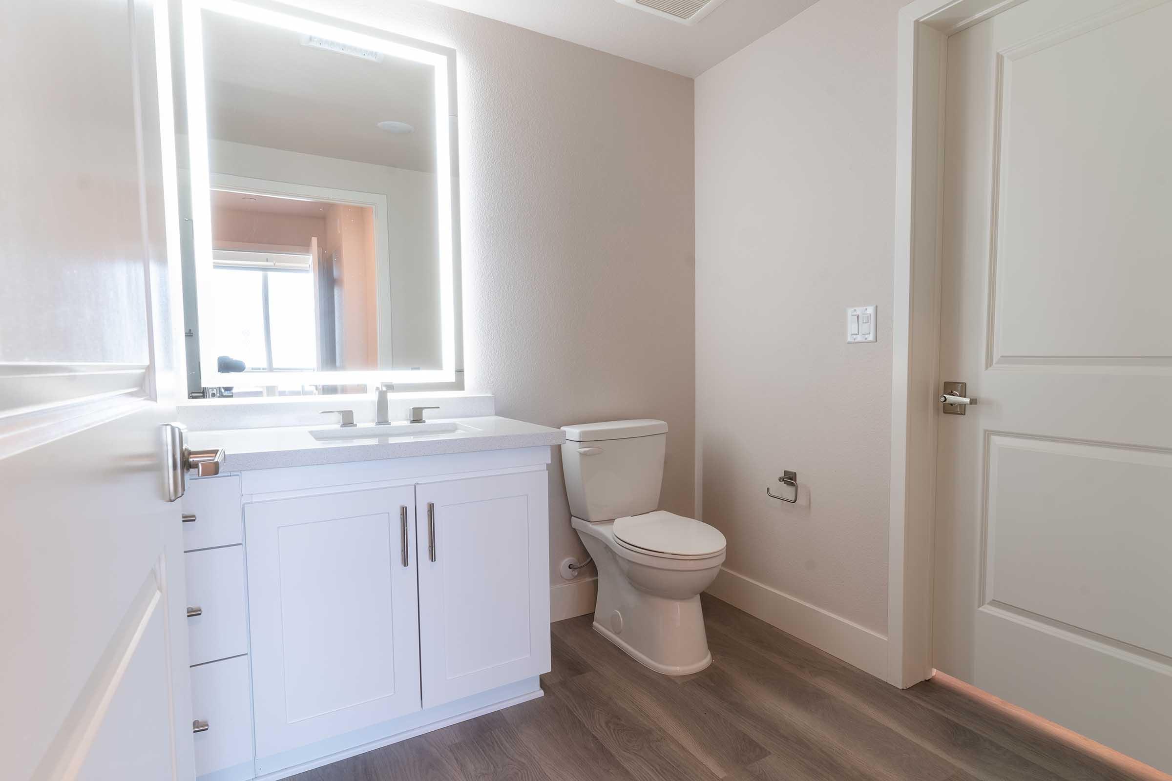 Modern bathroom featuring a white cabinet vanity with dual sinks, a large backlit mirror, and a toilet. The room has light-colored walls, a wooden floor, and a door leading elsewhere. Natural light is visible through a window in the background.