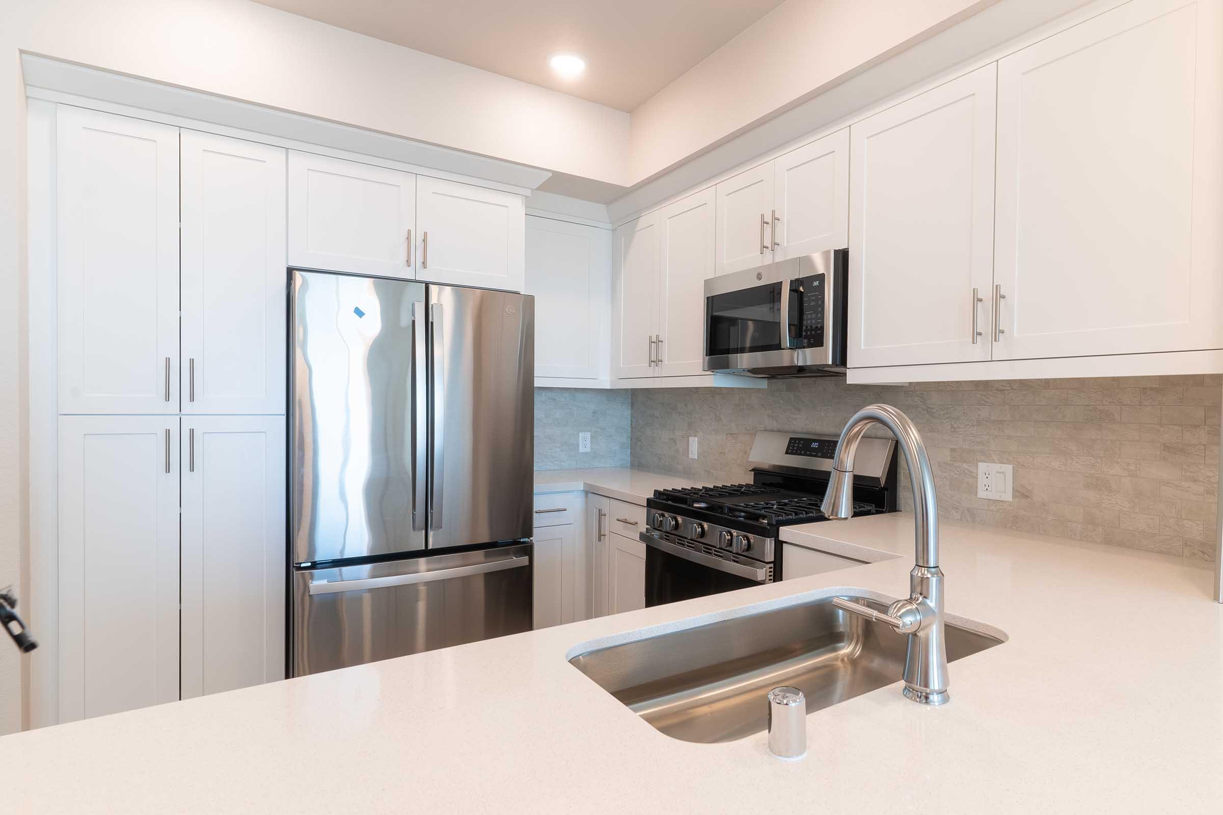 Modern kitchen featuring stainless steel appliances, including a refrigerator and microwave, with white cabinetry and a neutral stone backsplash. The sink is integrated into a light-colored countertop, and the design emphasizes a clean and contemporary aesthetic.