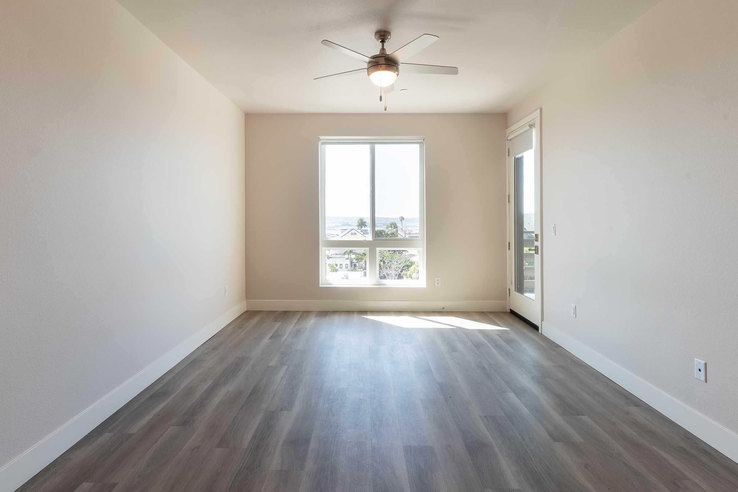 Bright, empty room with a light-colored wall and a large window allowing natural light to fill the space. The floor is covered in light wood-like vinyl, and there is a ceiling fan overhead. A glass door leads to an outdoor area, enhancing the open and airy feel of the room.