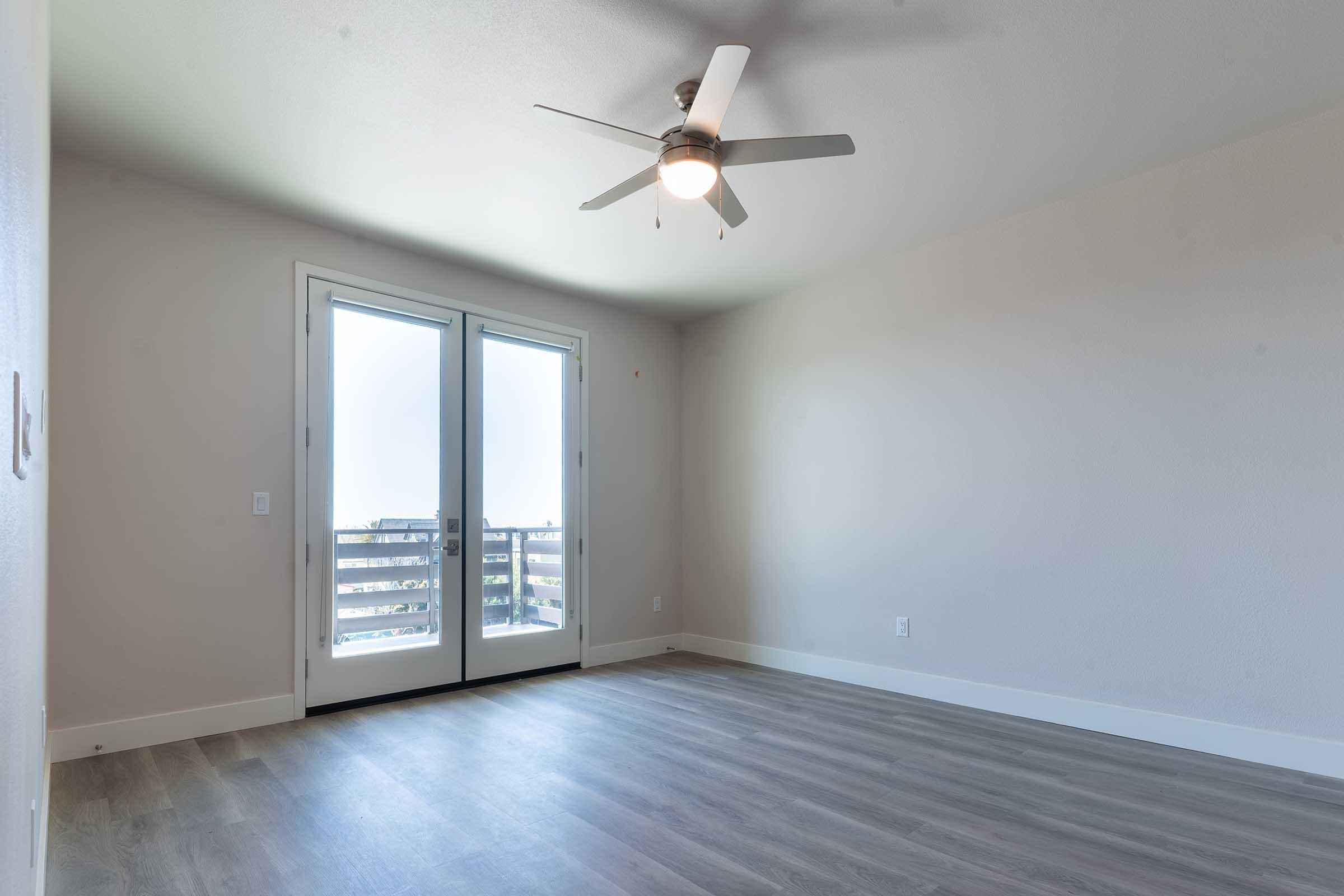 A spacious, empty room featuring light-colored walls, a ceiling fan, and large sliding glass doors that open to a balcony or outdoor area. The floor is made of light wood or laminate, allowing natural light to fill the space, creating a bright and airy atmosphere.