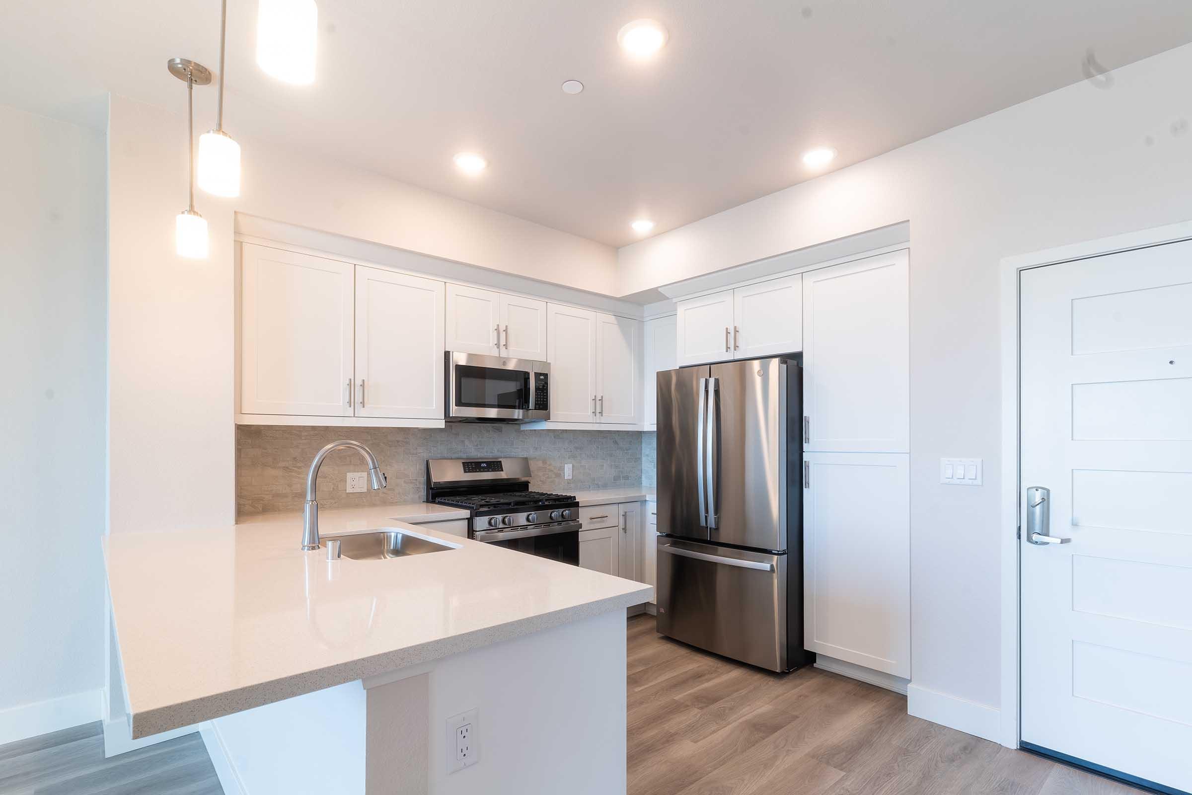 Modern kitchen featuring white cabinets, stainless steel appliances including a microwave and refrigerator, and a gas stove. The countertop is light-colored quartz, and there are pendant lights above the island. A door with a simple design is visible, leading to the outside. Bright and inviting space with a contemporary feel.