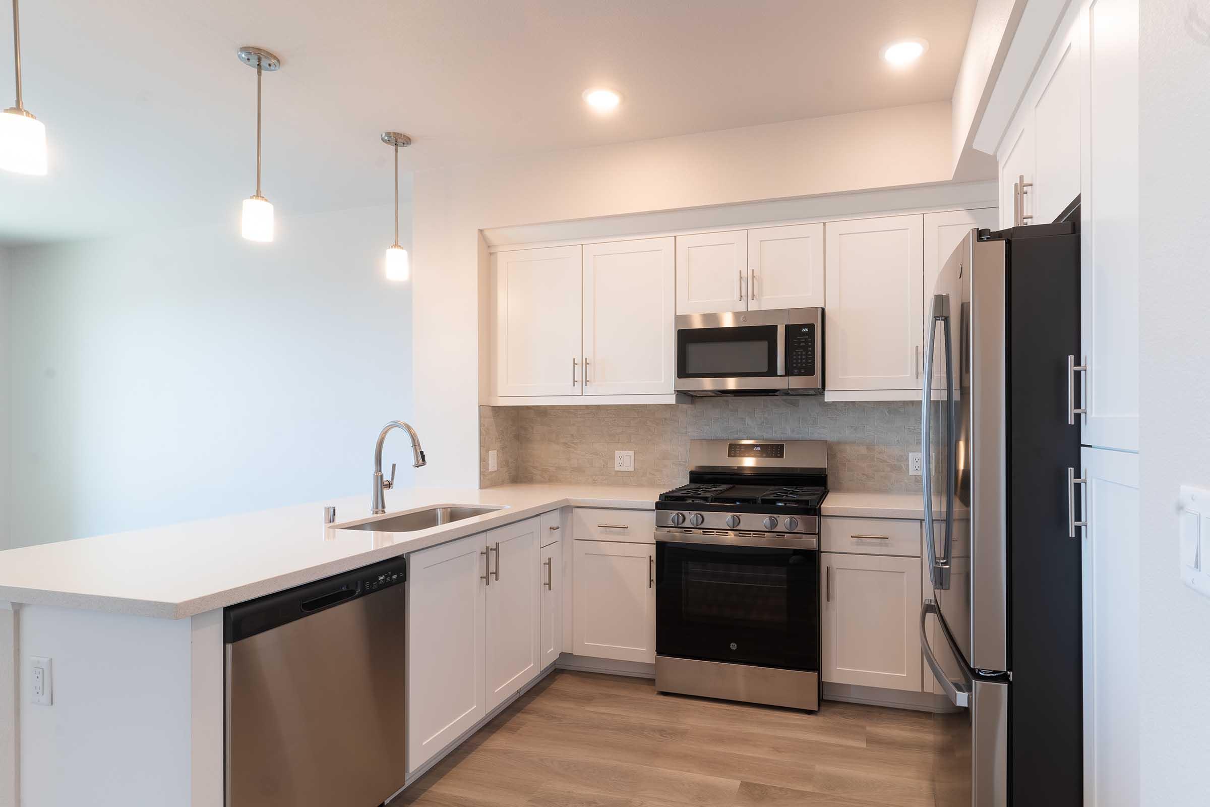 Modern kitchen featuring white cabinetry, stainless steel appliances including a microwave and gas stove, and a dishwasher. The space has a gray backsplash, bright lighting from pendant fixtures, and light wood flooring, creating a bright and clean aesthetic.