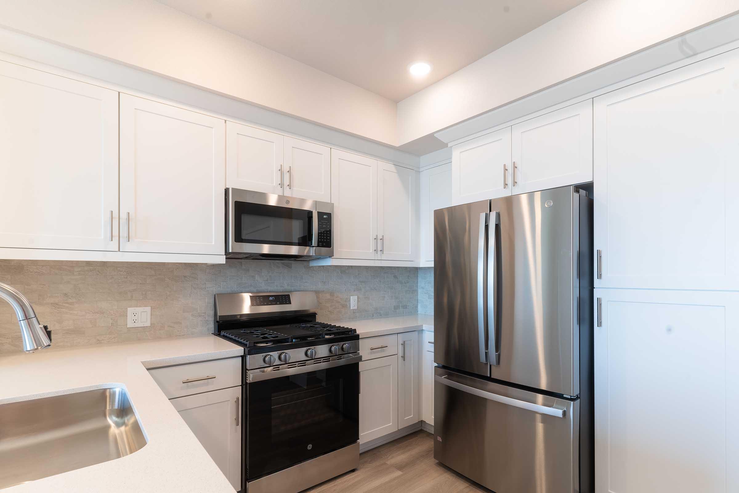 Modern kitchen featuring white cabinetry, stainless steel appliances including a gas stove, microwave, and refrigerator. The countertop is light-colored, and there is a sink with a sleek faucet. The walls are painted light, creating a bright and airy ambiance.