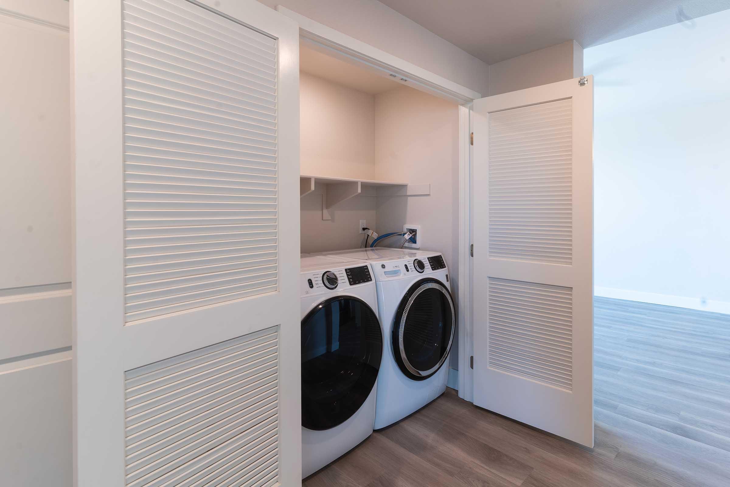 A laundry area featuring a pair of front-loading washing machines with a shelf above for storage, enclosed by white louvered doors. The room has light-colored walls and hardwood-style flooring, creating a modern and clean look.